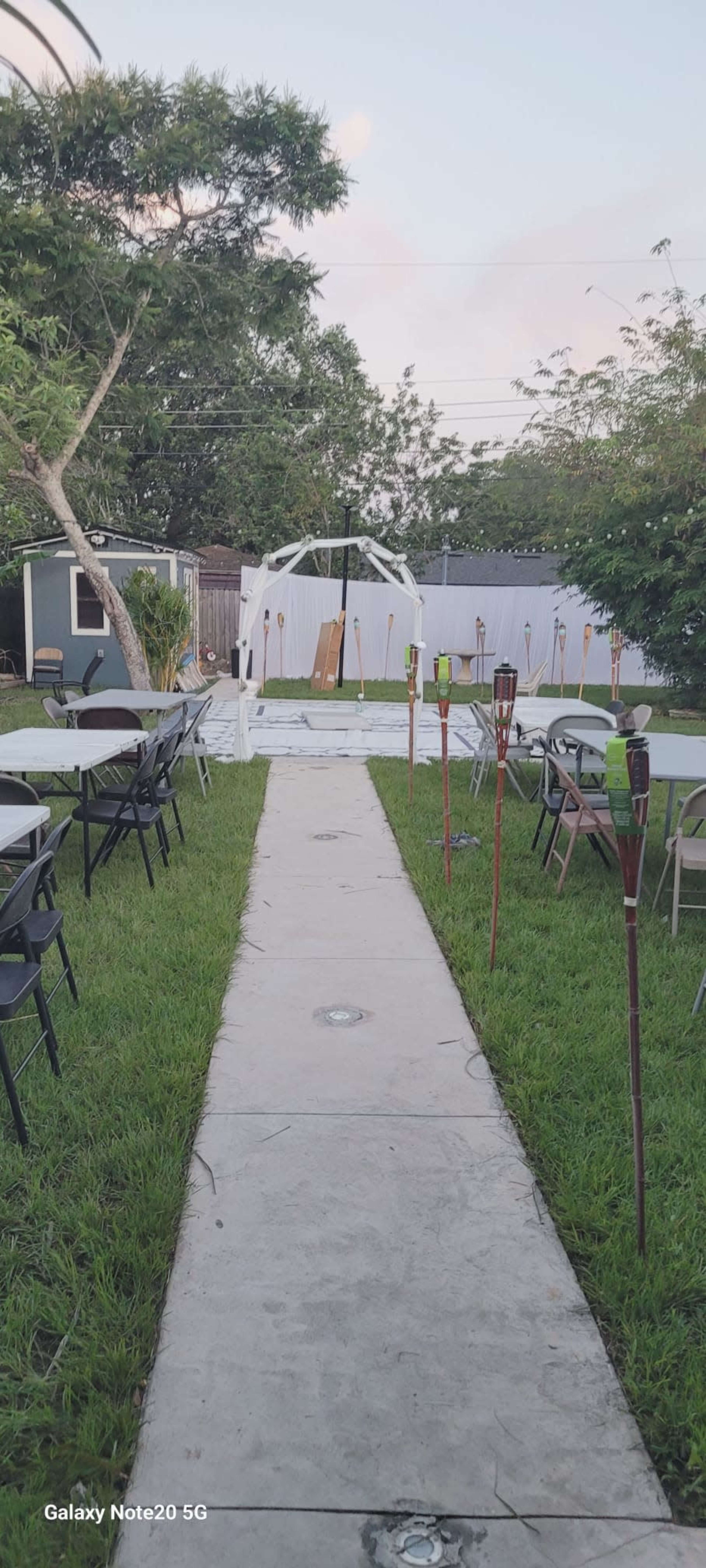The image shows a pathway leading to a tent set up in a grassy area, flanked by chairs and tiki torches.