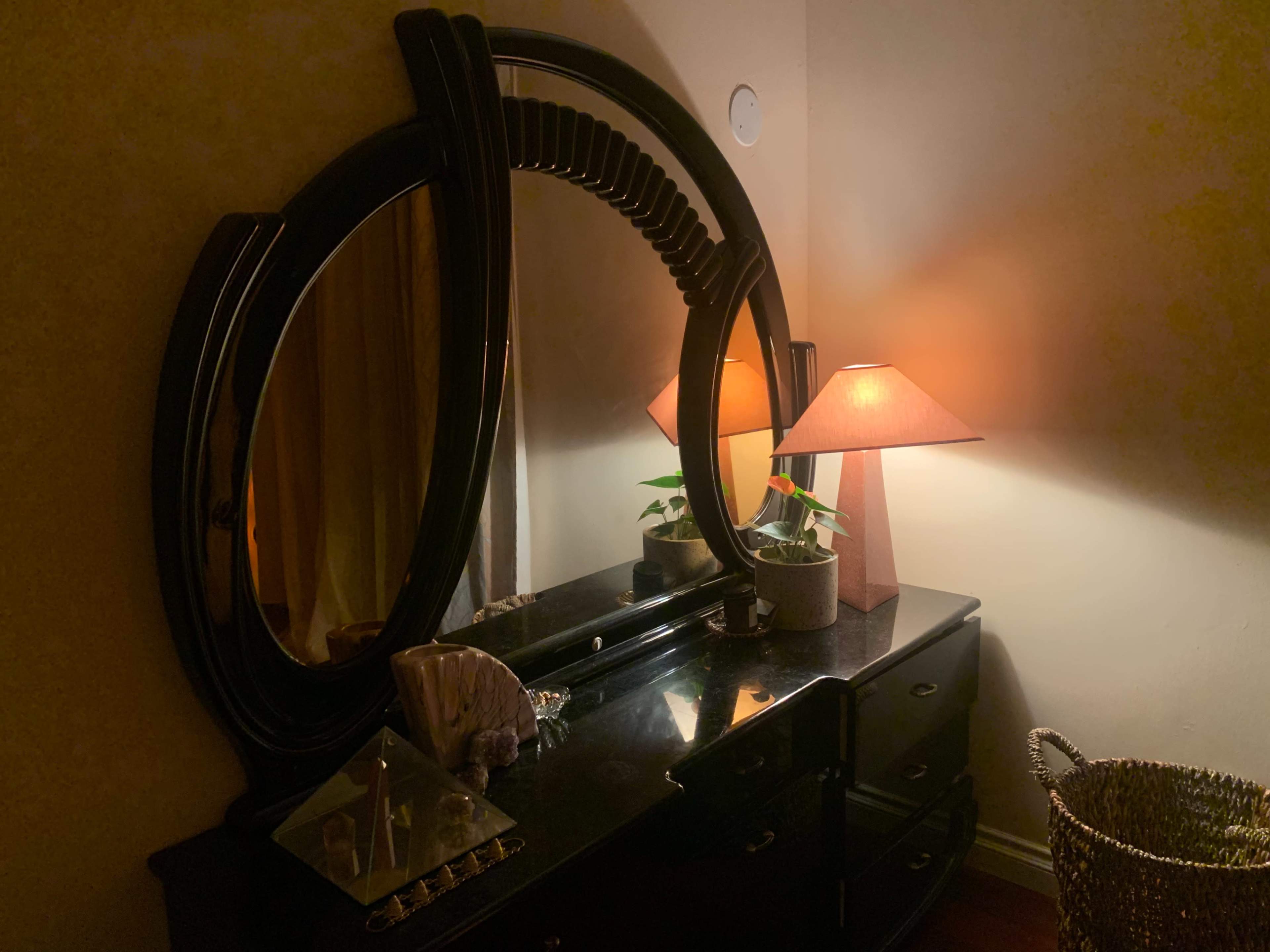 A black dresser with a round mirror and a lamp casting a warm light, accompanied by a potted plant and a decorative dish.