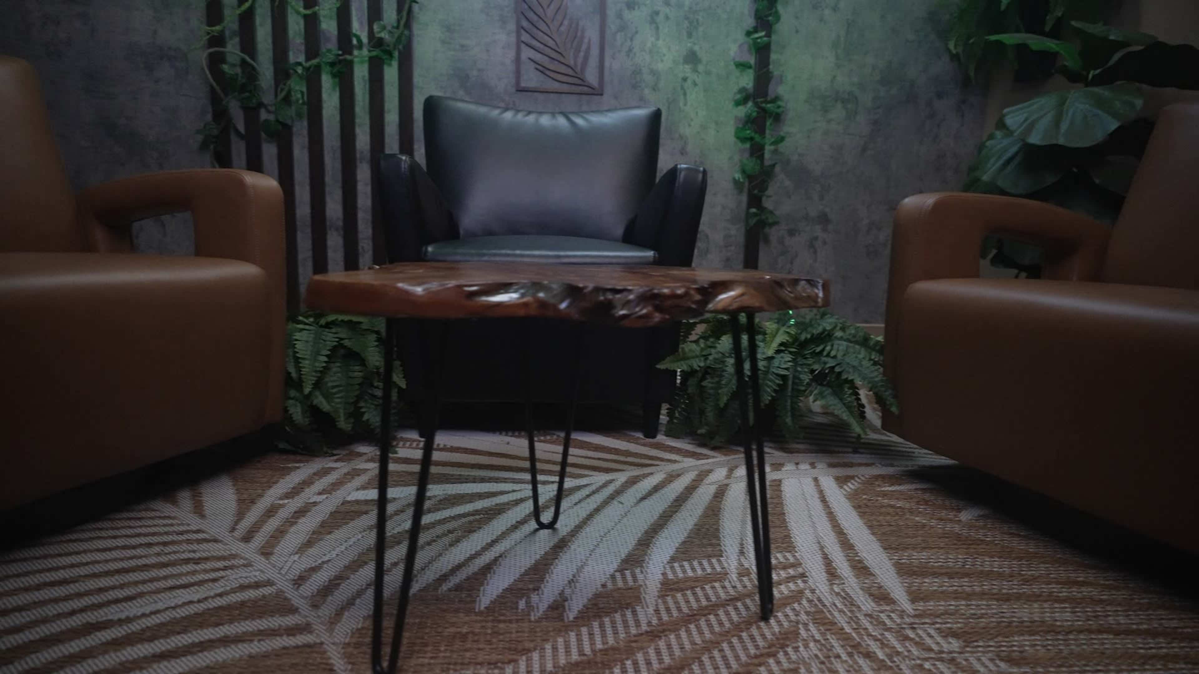 The image shows a small wooden table with black legs positioned between two brown armchairs in a room with greenery and textured walls.