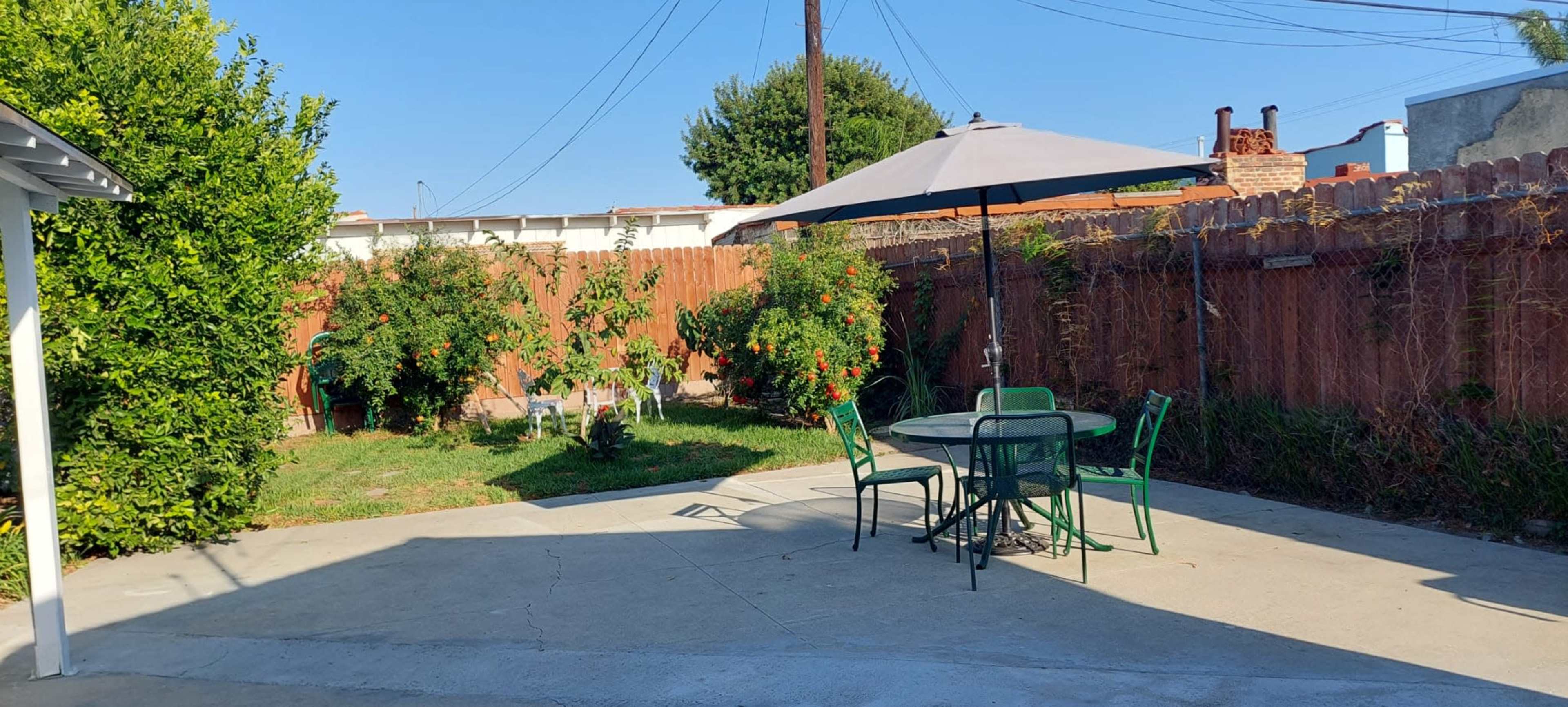 The image shows a backyard with a round table and chairs under a gray umbrella, surrounded by green grass and plants.