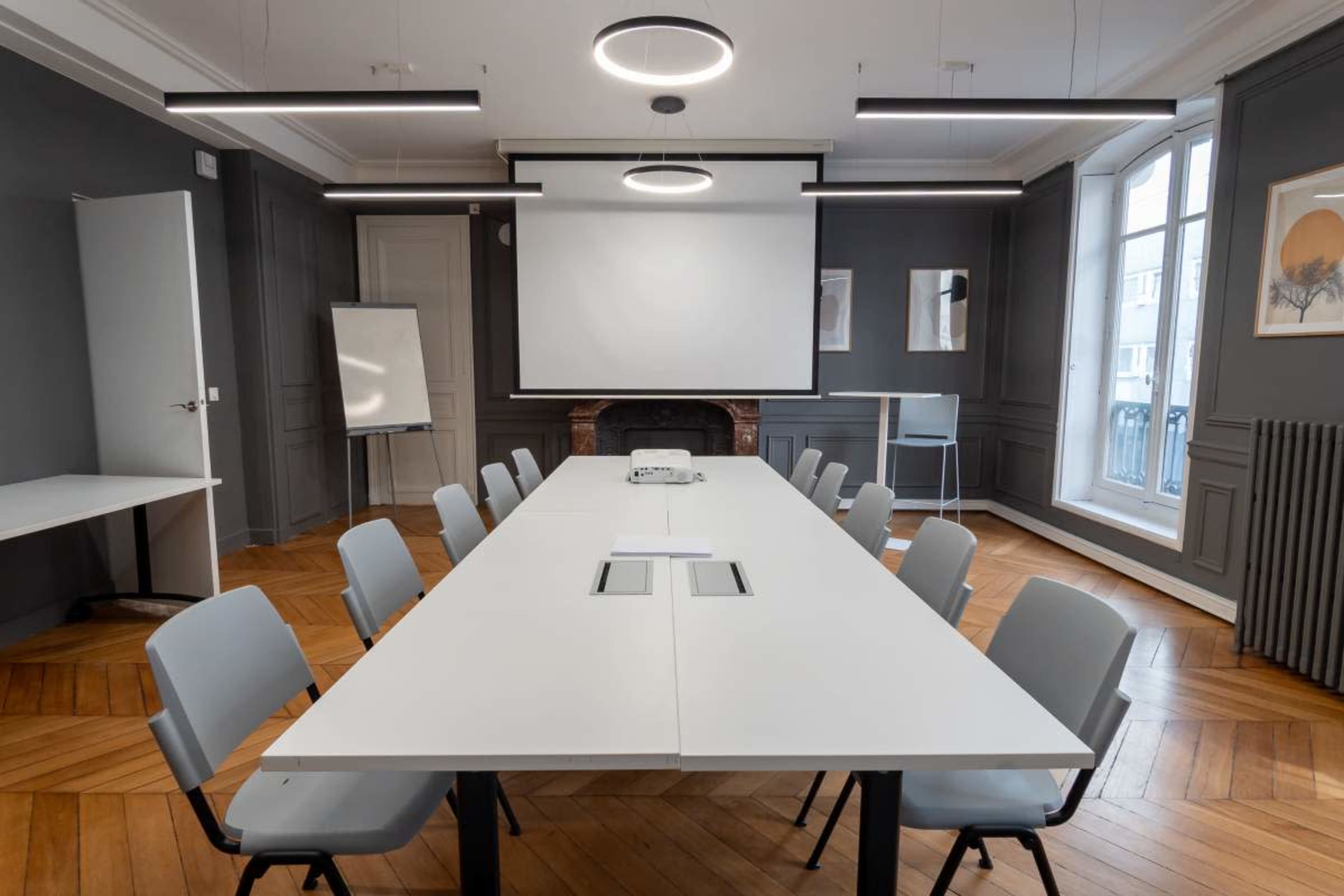 A modern conference room features a long table surrounded by gray chairs, with a large screen at the front and large windows providing natural light.