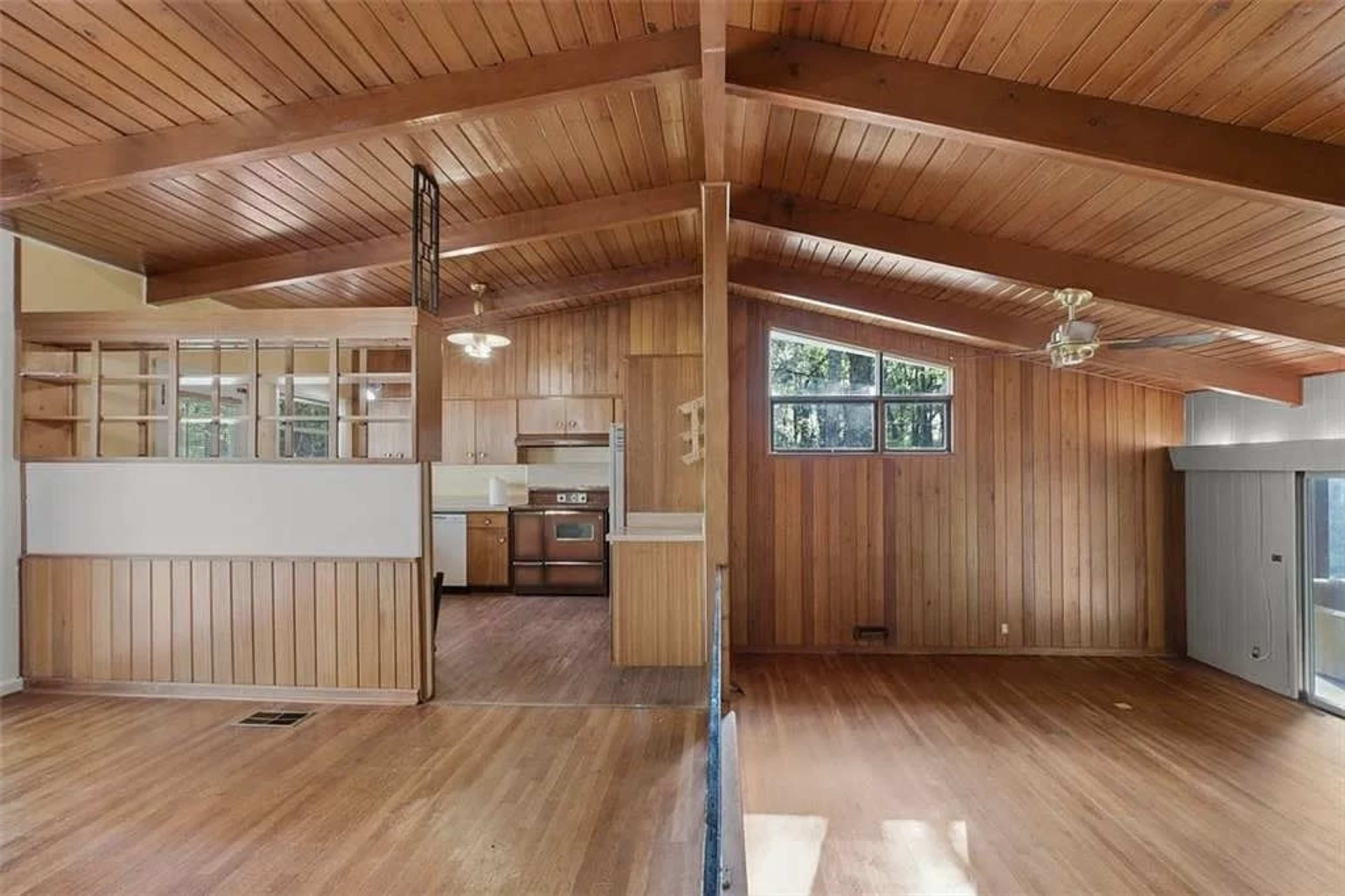 The image shows a spacious interior of a house featuring high wooden beams, a kitchen area with appliances, and large windows allowing natural light.