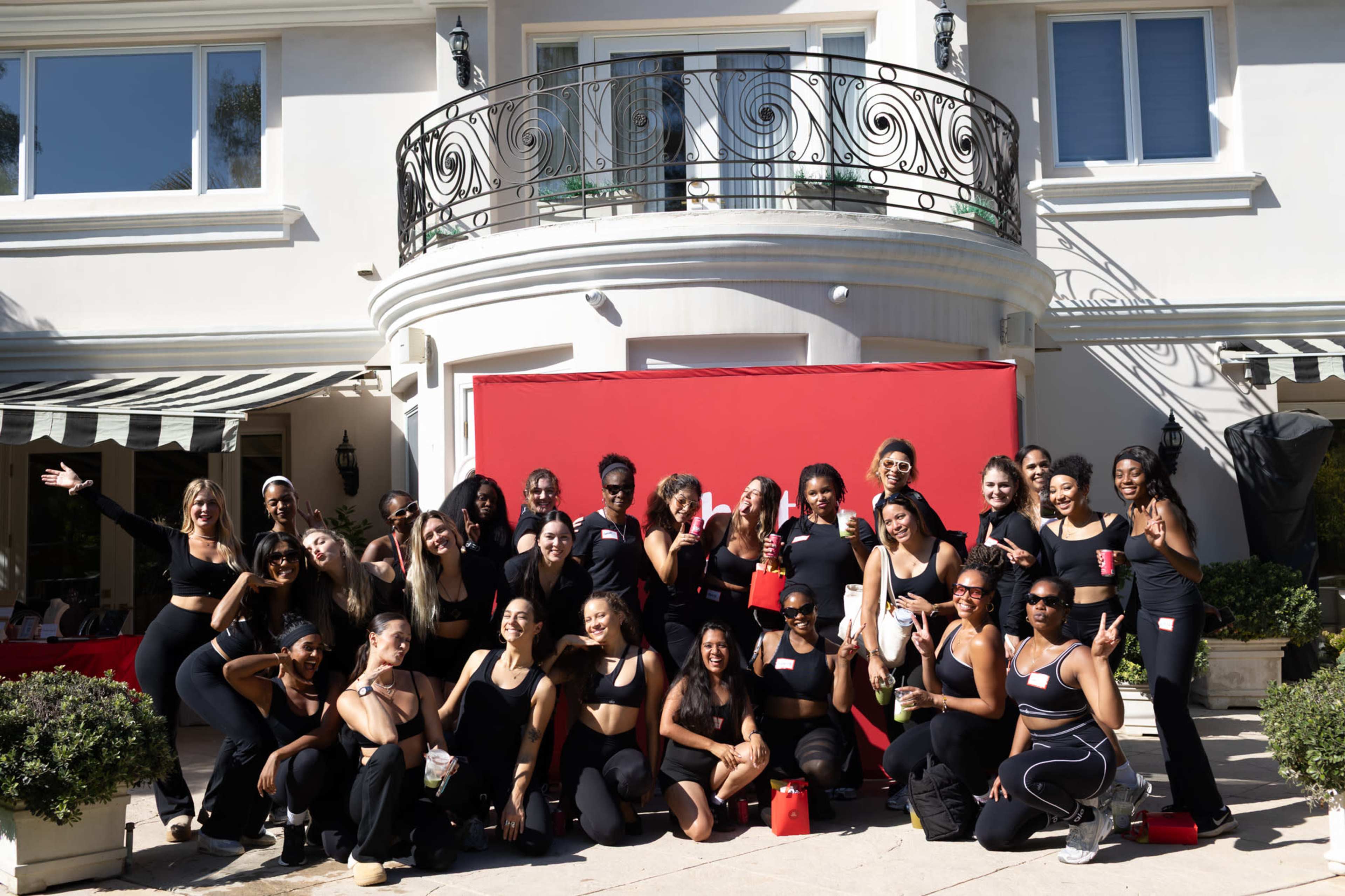 A large group of women, dressed in matching black outfits, poses together in front of a red backdrop outside a building with a balcony.