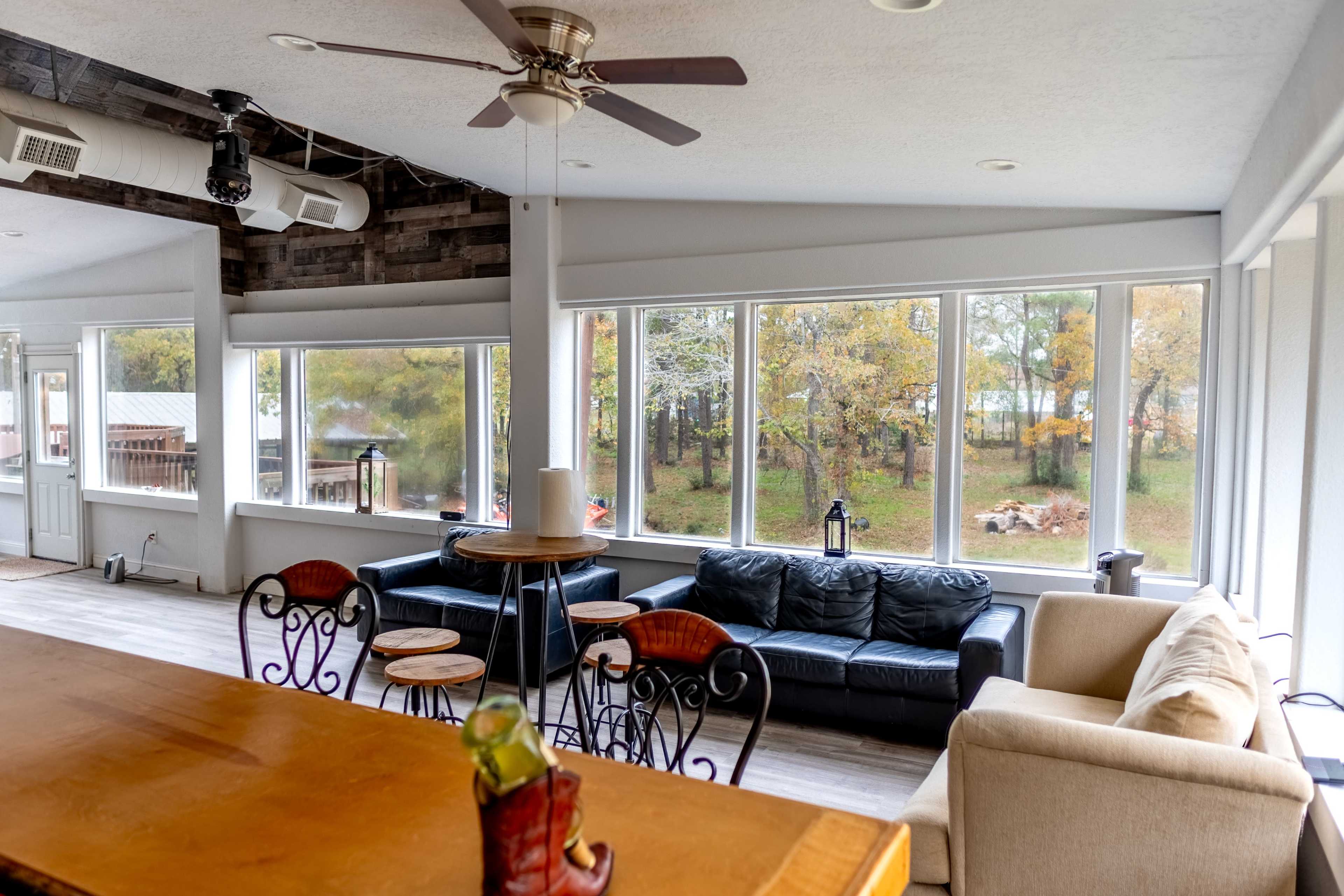 The image shows a modern interior living space with large windows, a mix of seating options, and a wooden dining table.