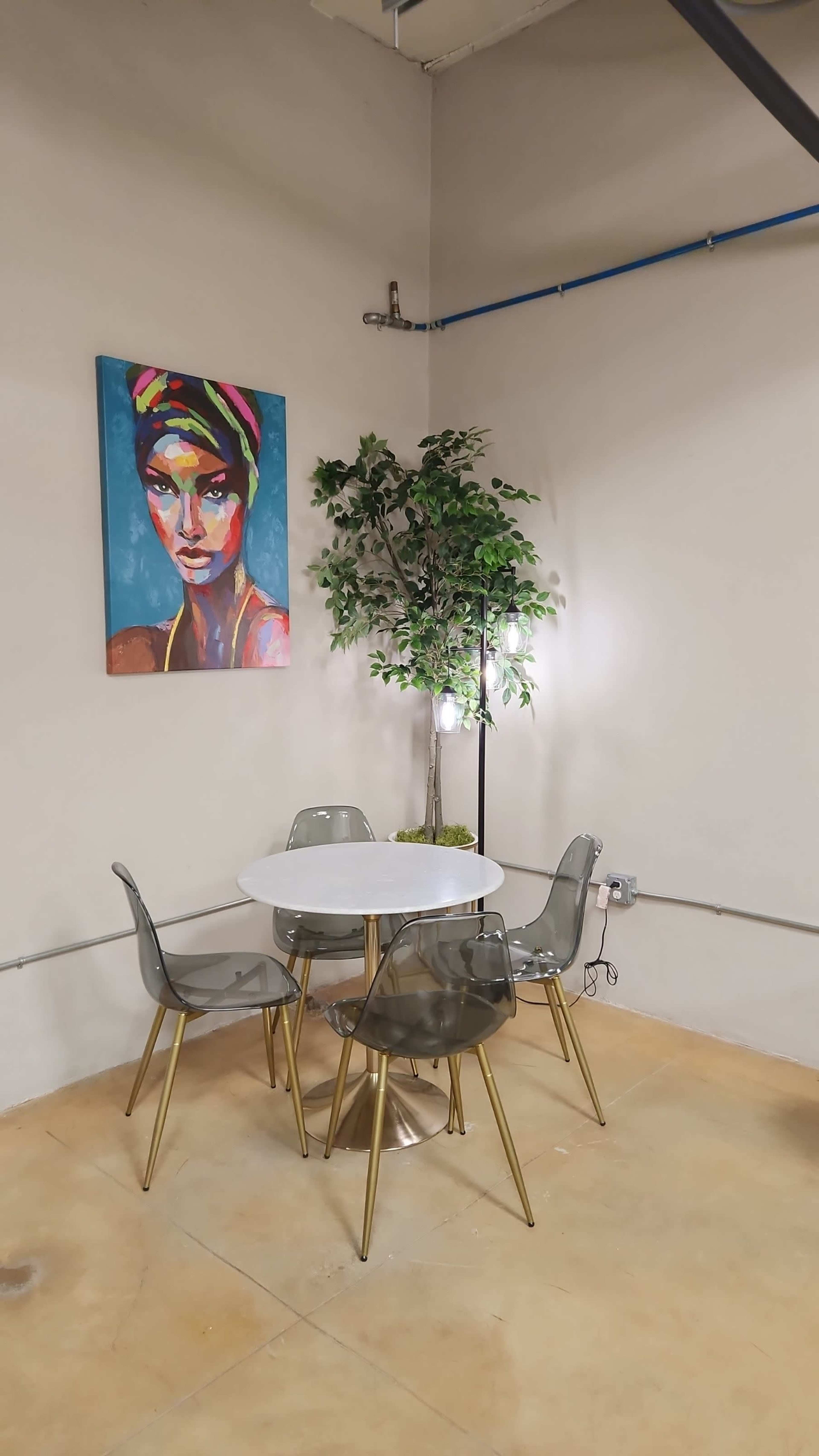 A small dining area features a round white table surrounded by transparent chairs, with a colorful portrait on the wall and a potted plant nearby.