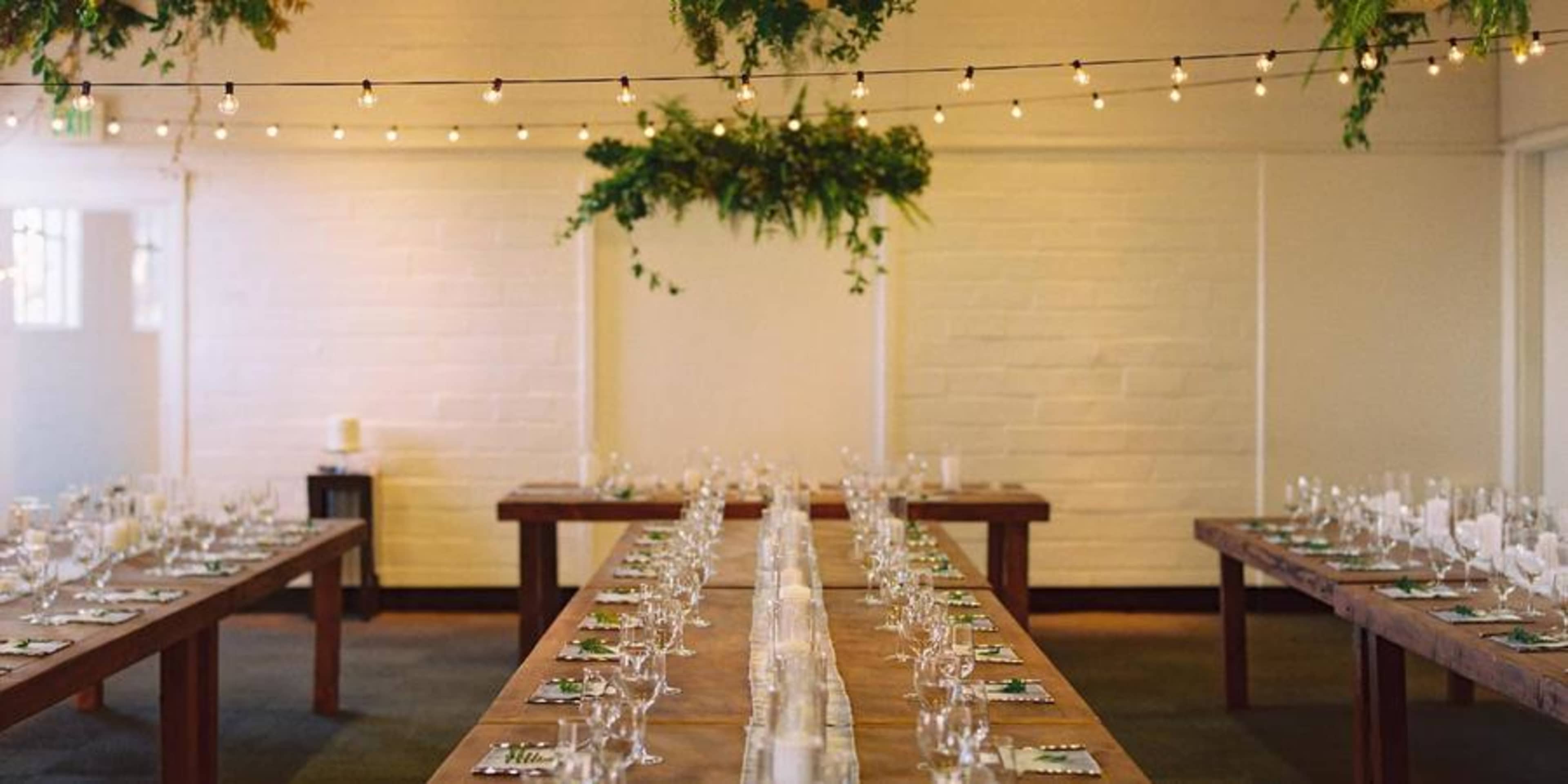 A long dining table is set with glassware and candles under decorations of greenery and string lights.