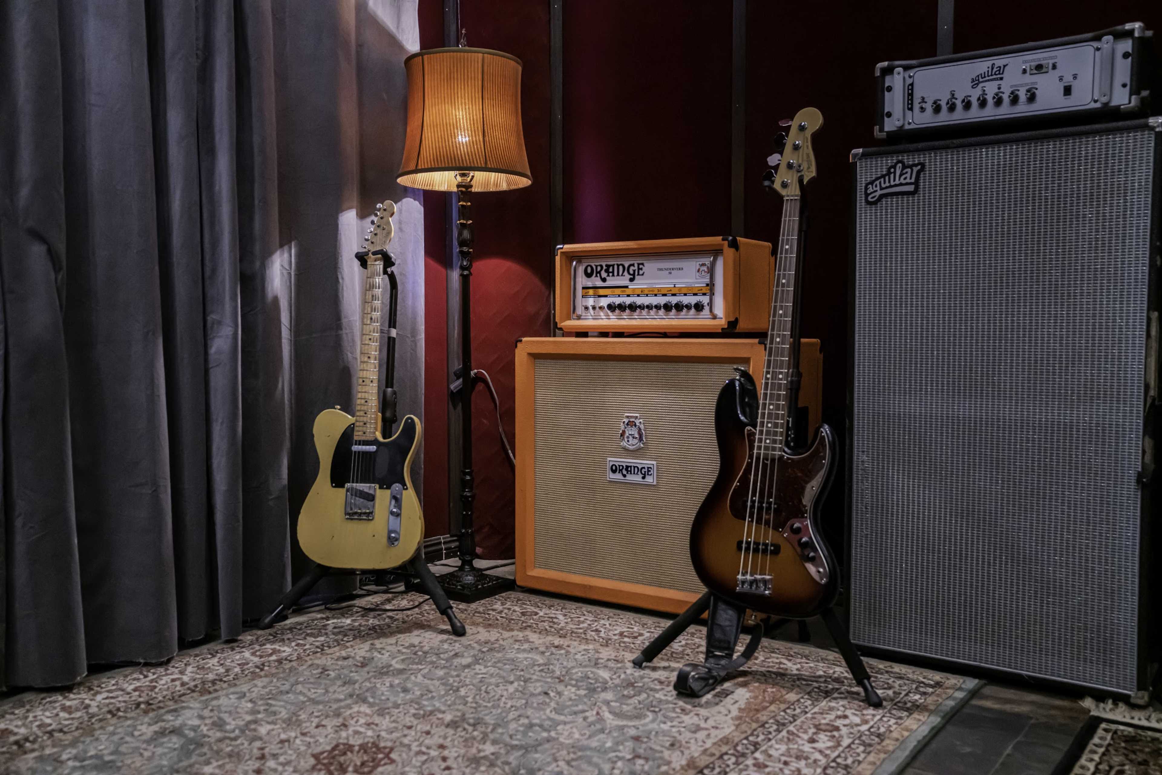 The scene features a guitar and a bass leaning against an orange amplifier and a patterned rug, with a lamp casting light in the corner of a room.