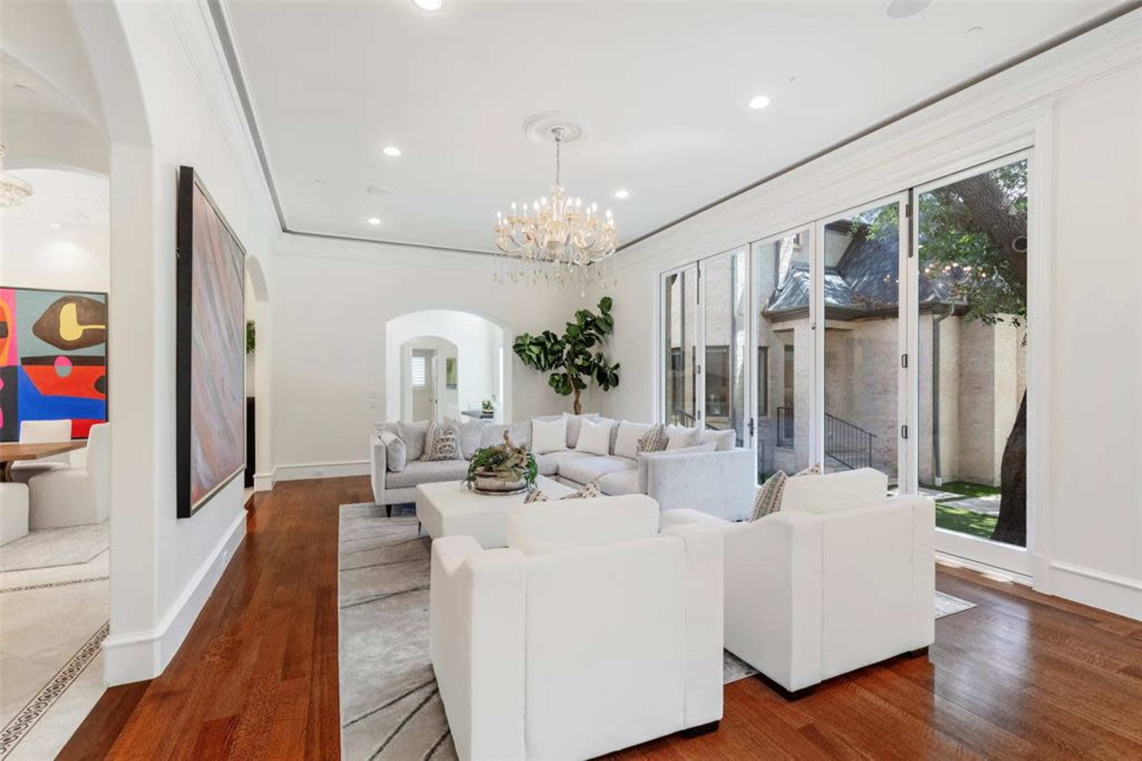 The image shows a bright living room with white furniture, large windows, and a chandelier, featuring a view of a garden through the glass doors.