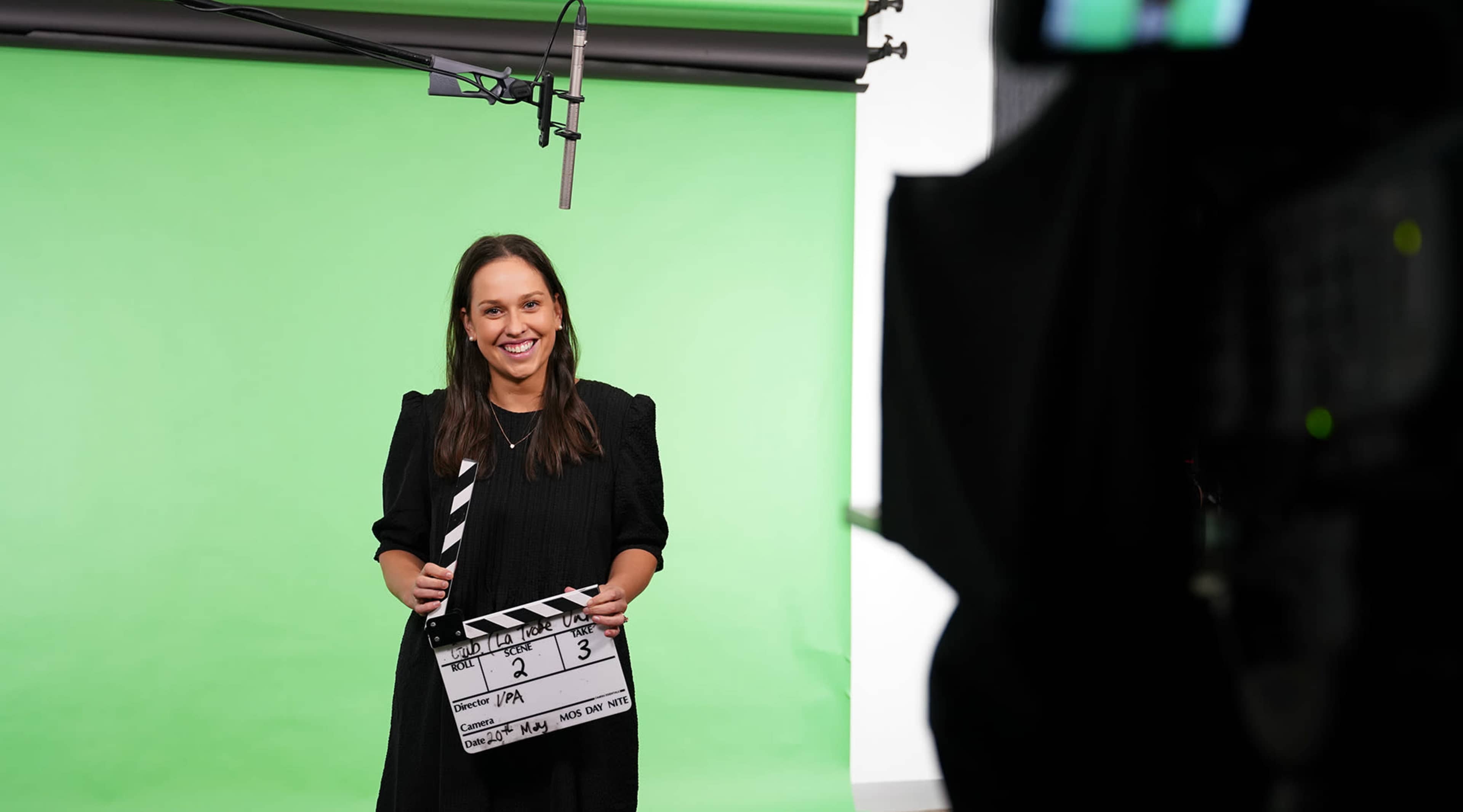 A person holds a clapperboard while standing in front of a green screen in a studio setting.