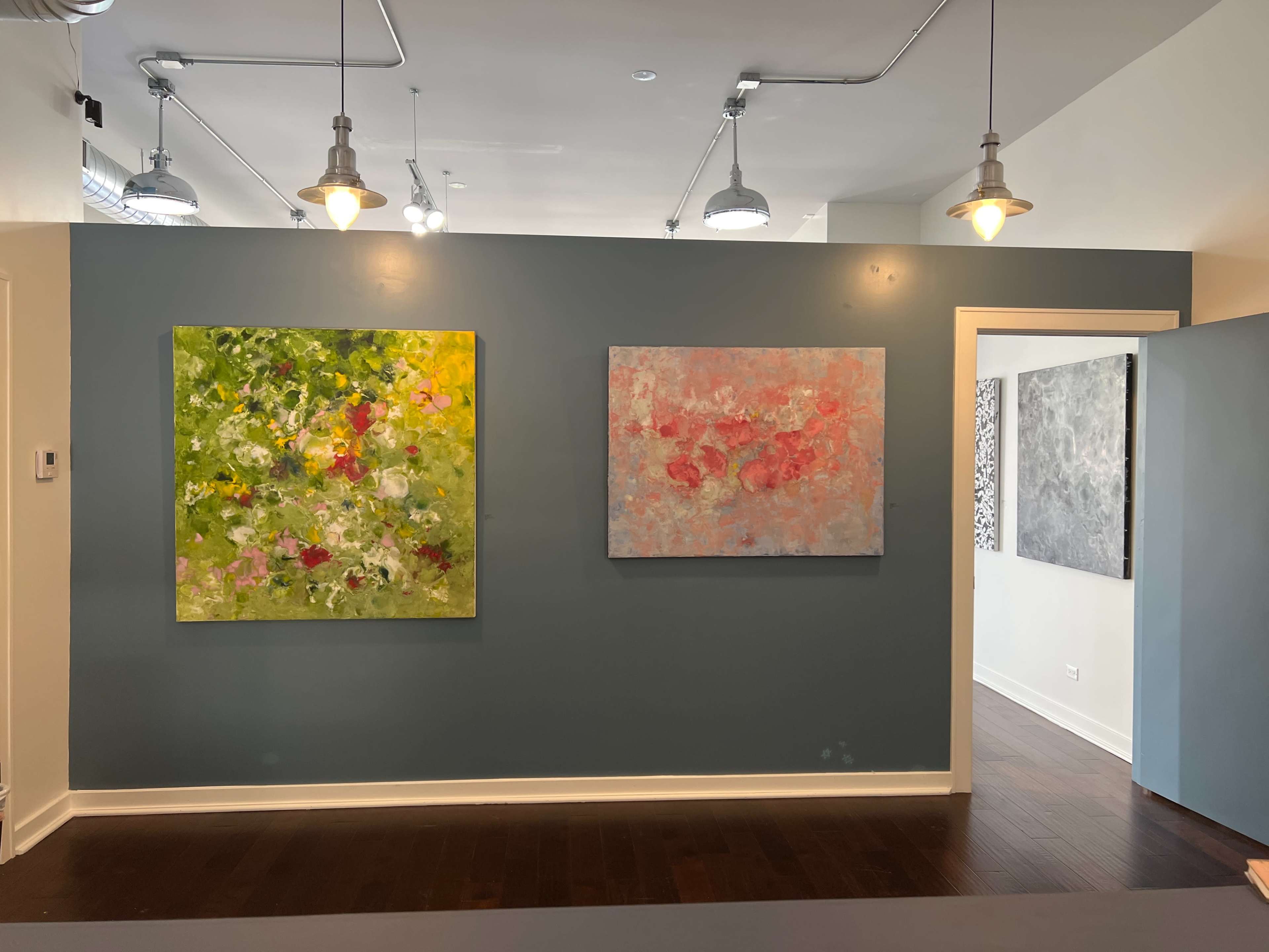 A brightly lit room featuring three abstract paintings on a teal wall, with pendant lights and wooden flooring.