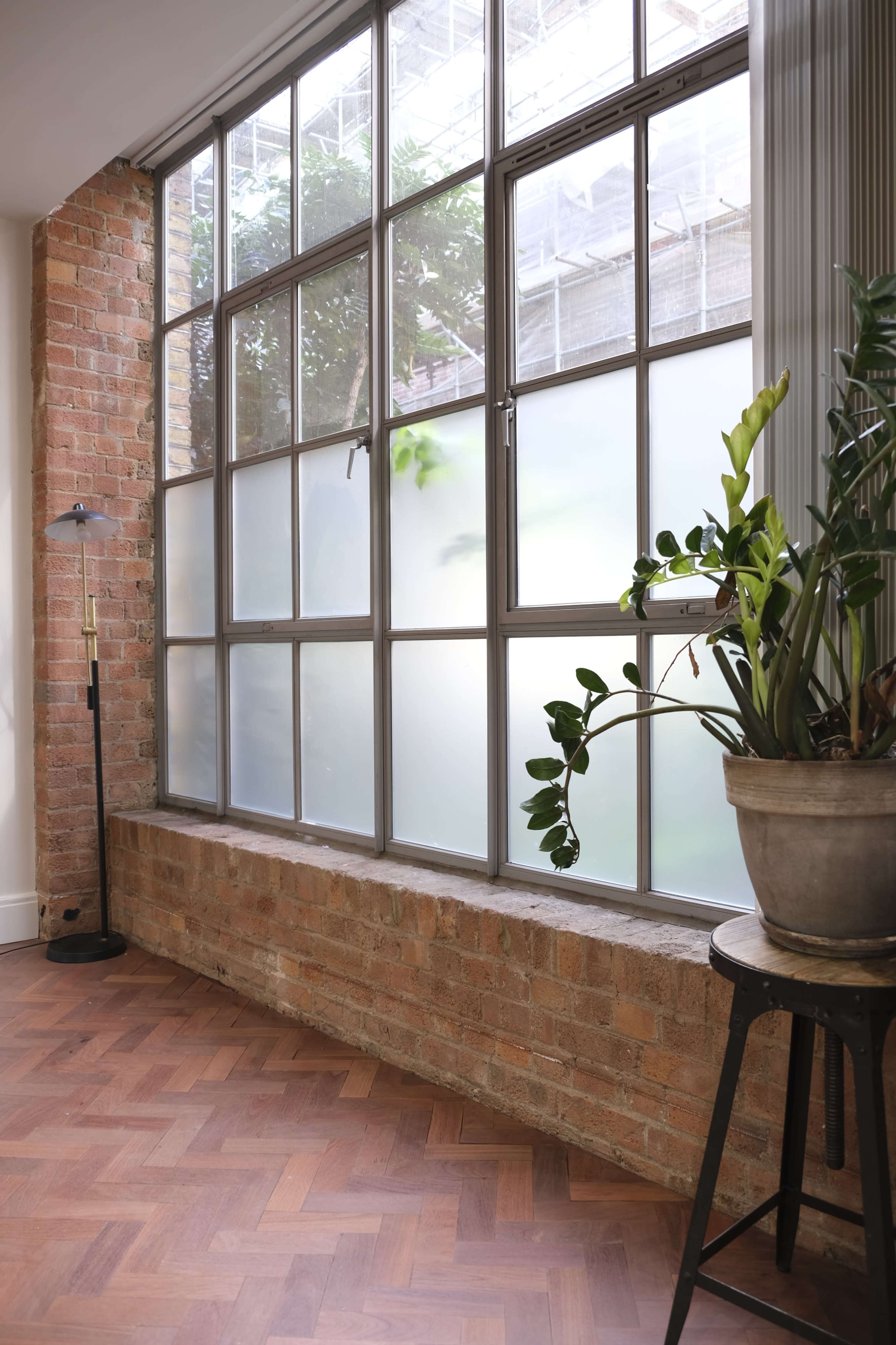 A room with a large window featuring metal frames, a brick wall, a potted plant, and a small table against a wooden floor.