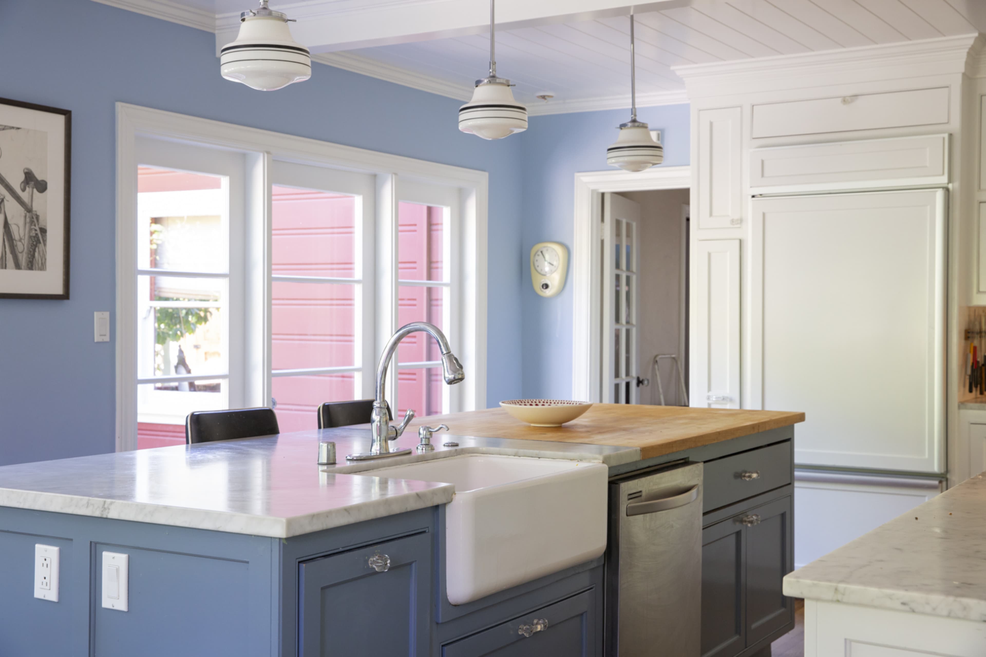 A kitchen features blue walls, a central island with a marble countertop and farmhouse sink, and large windows allowing natural light.