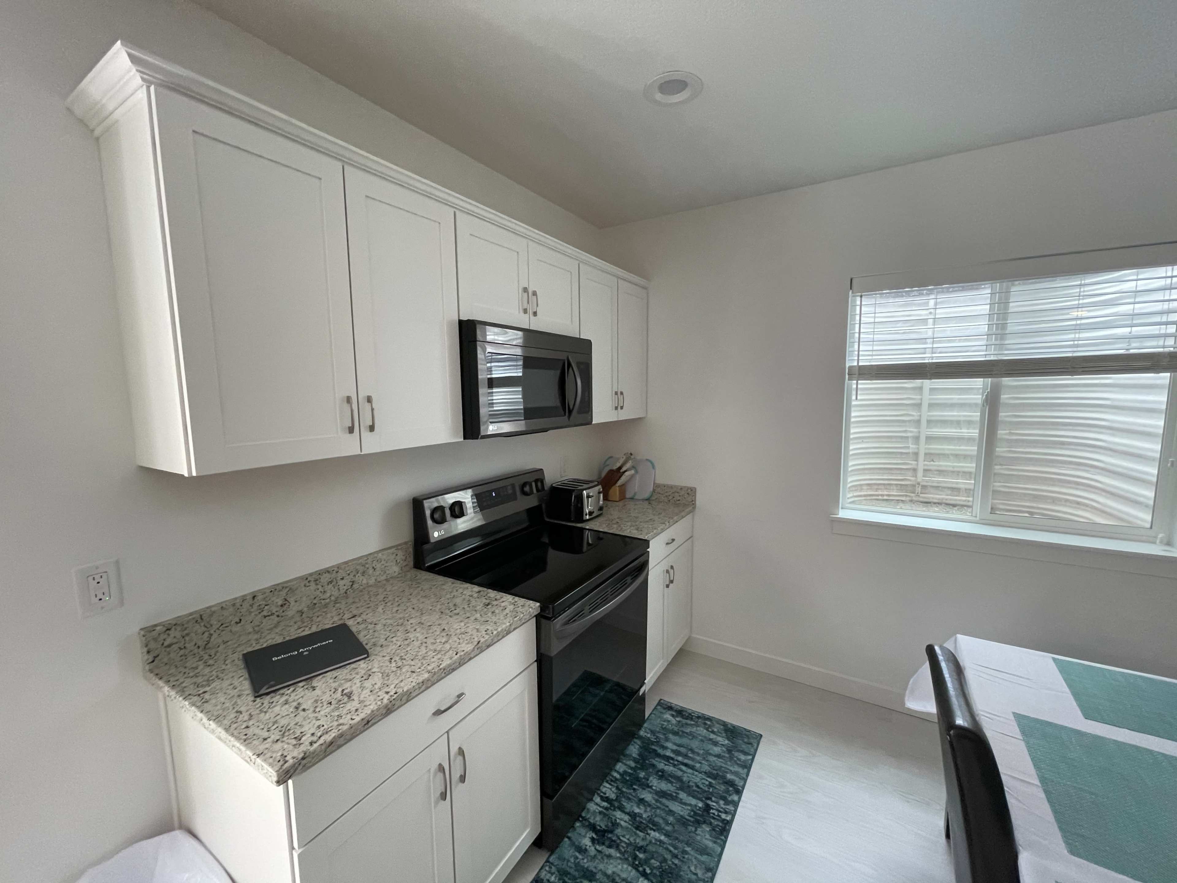 A modern kitchen featuring white cabinets, a black microwave and stove, and a window with blinds overlooking a light-colored wall.