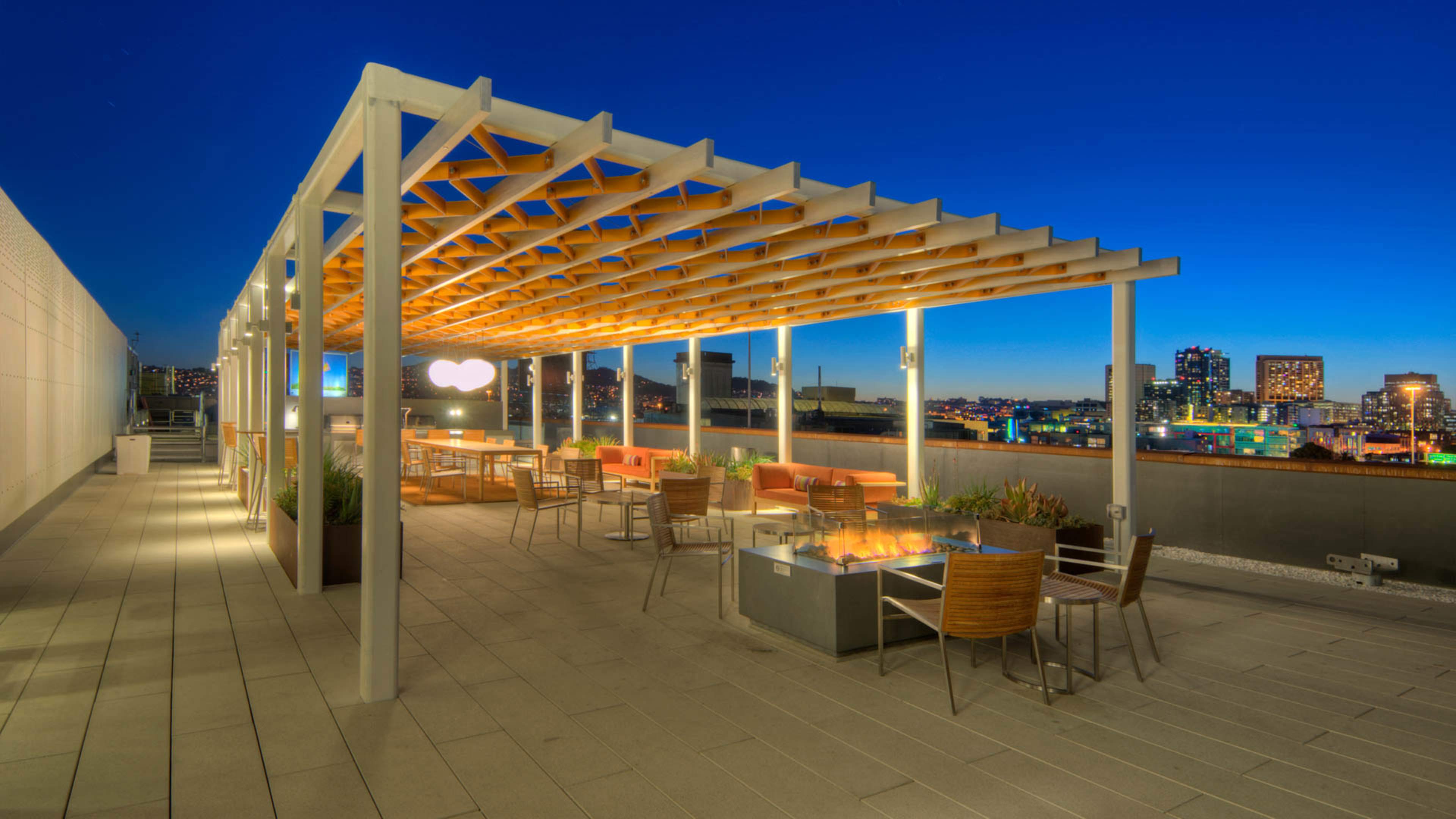 A rooftop terrace with a pergola, seating arrangements, and a fire pit against a twilight city skyline.