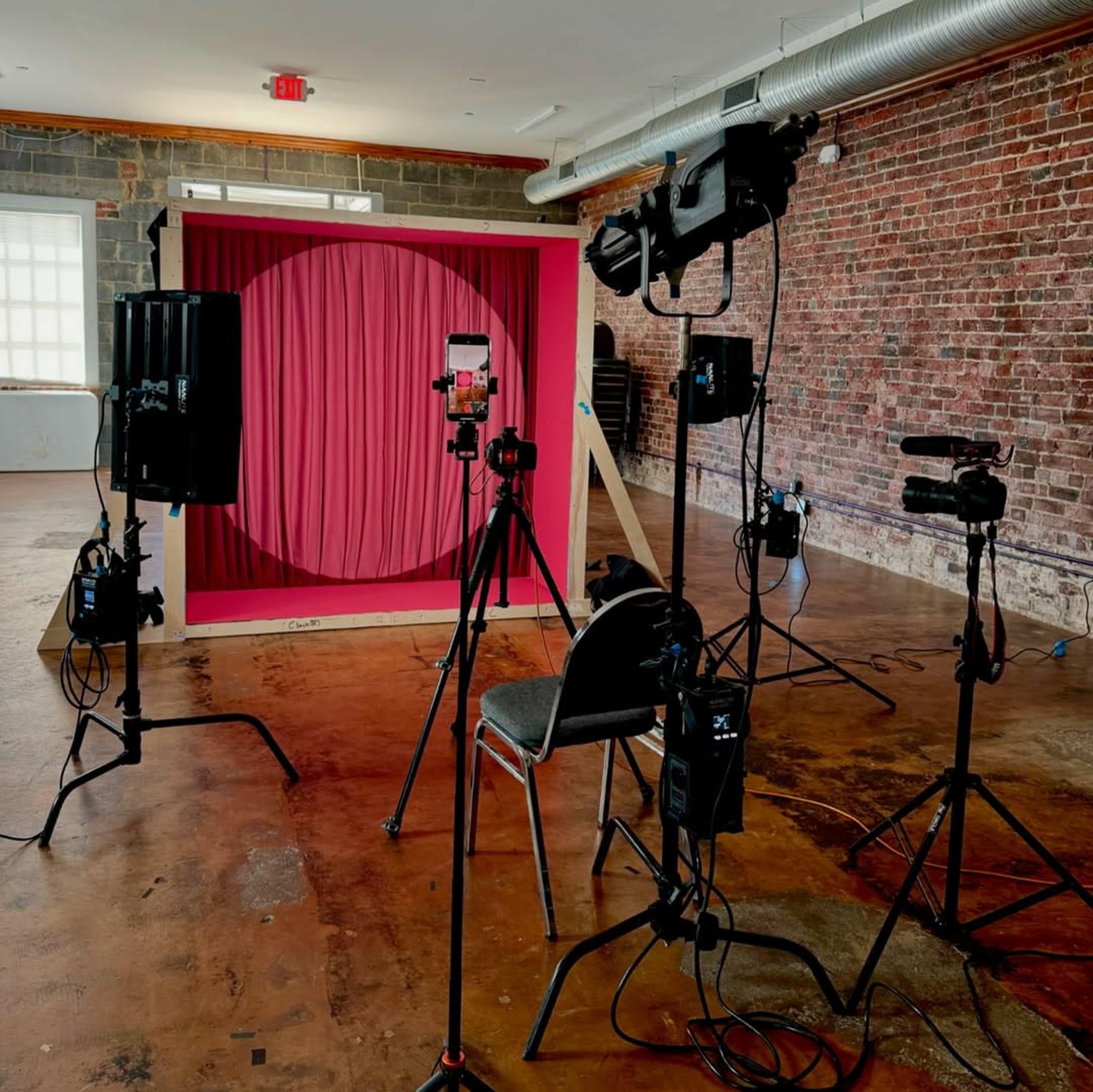The image shows a photography setup in a room featuring a pink backdrop, several cameras on tripods, and studio lights arranged around a chair.