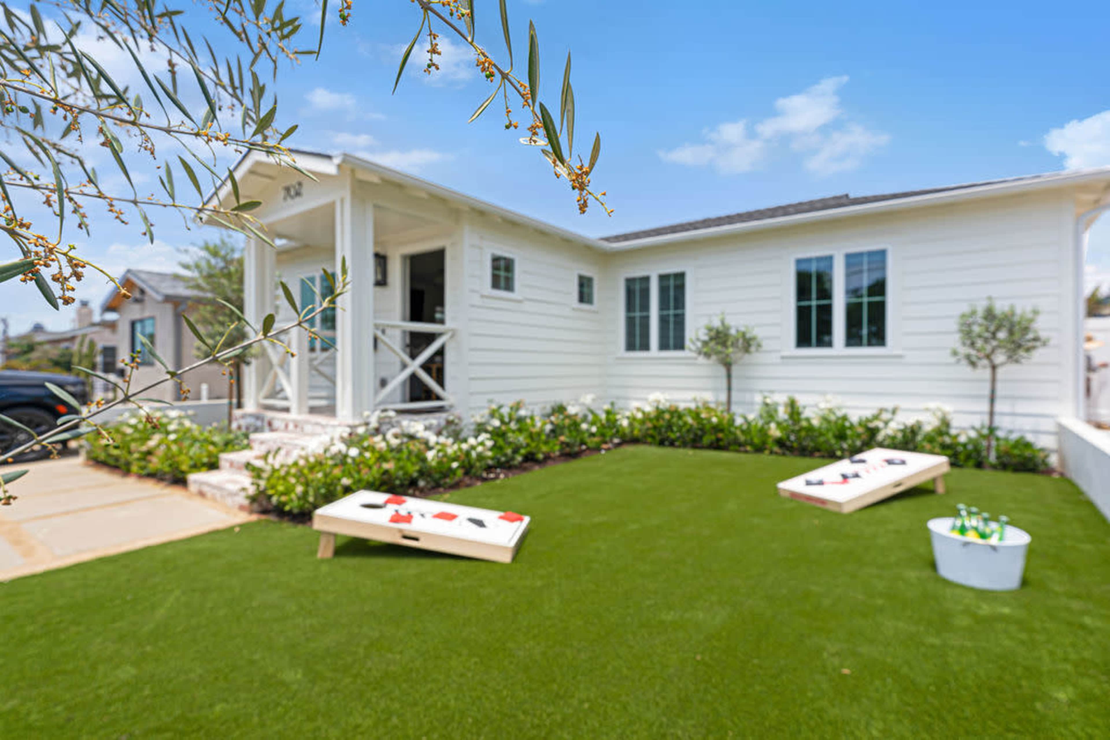 A single-story white house with a manicured lawn features two cornhole boards in the foreground and flowering bushes lining the driveway.