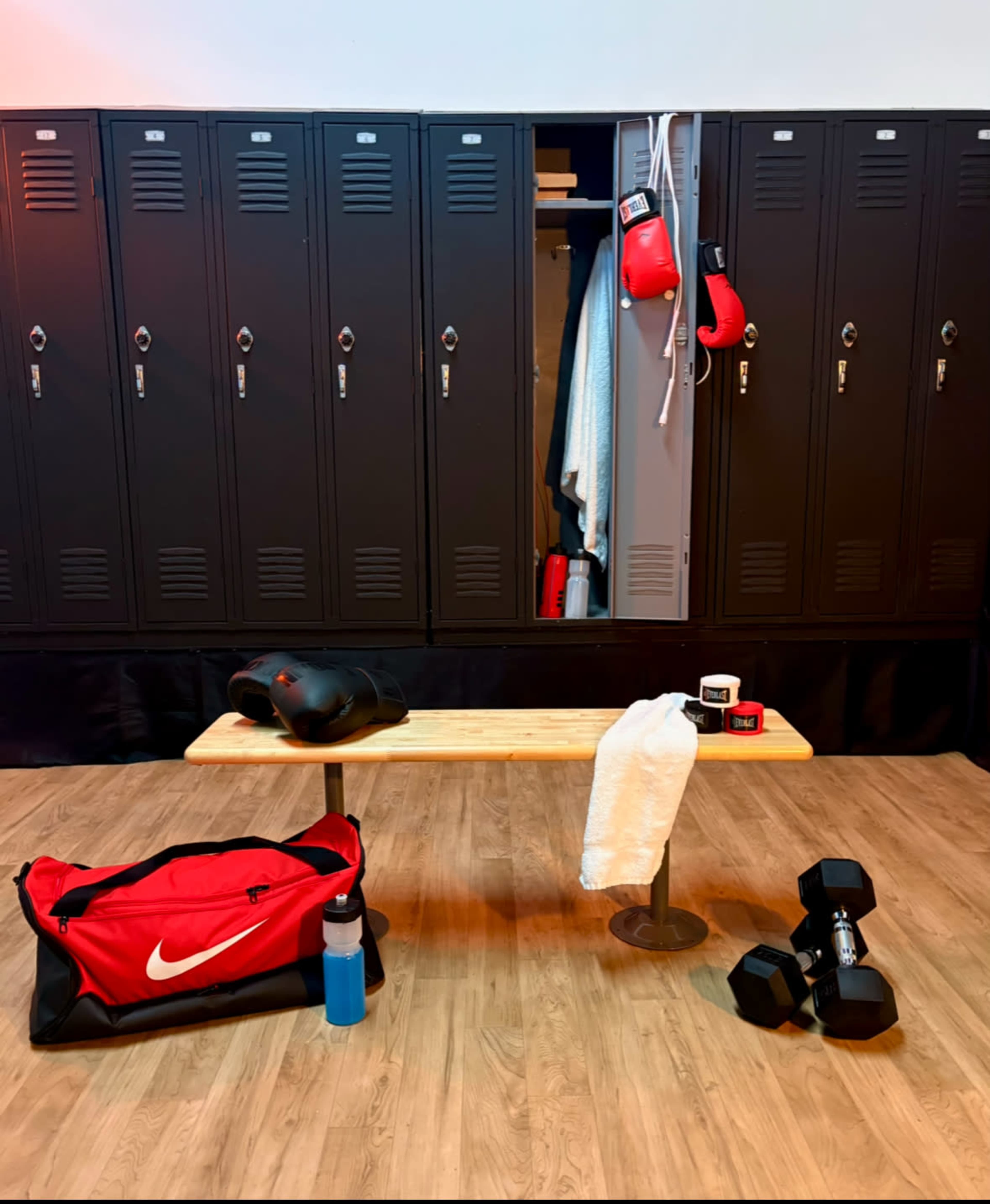 The image shows a gym locker room with several black lockers, a wooden bench, and workout gear including a red duffel bag, a water bottle, boxing gloves, a towel, and dumbbells.