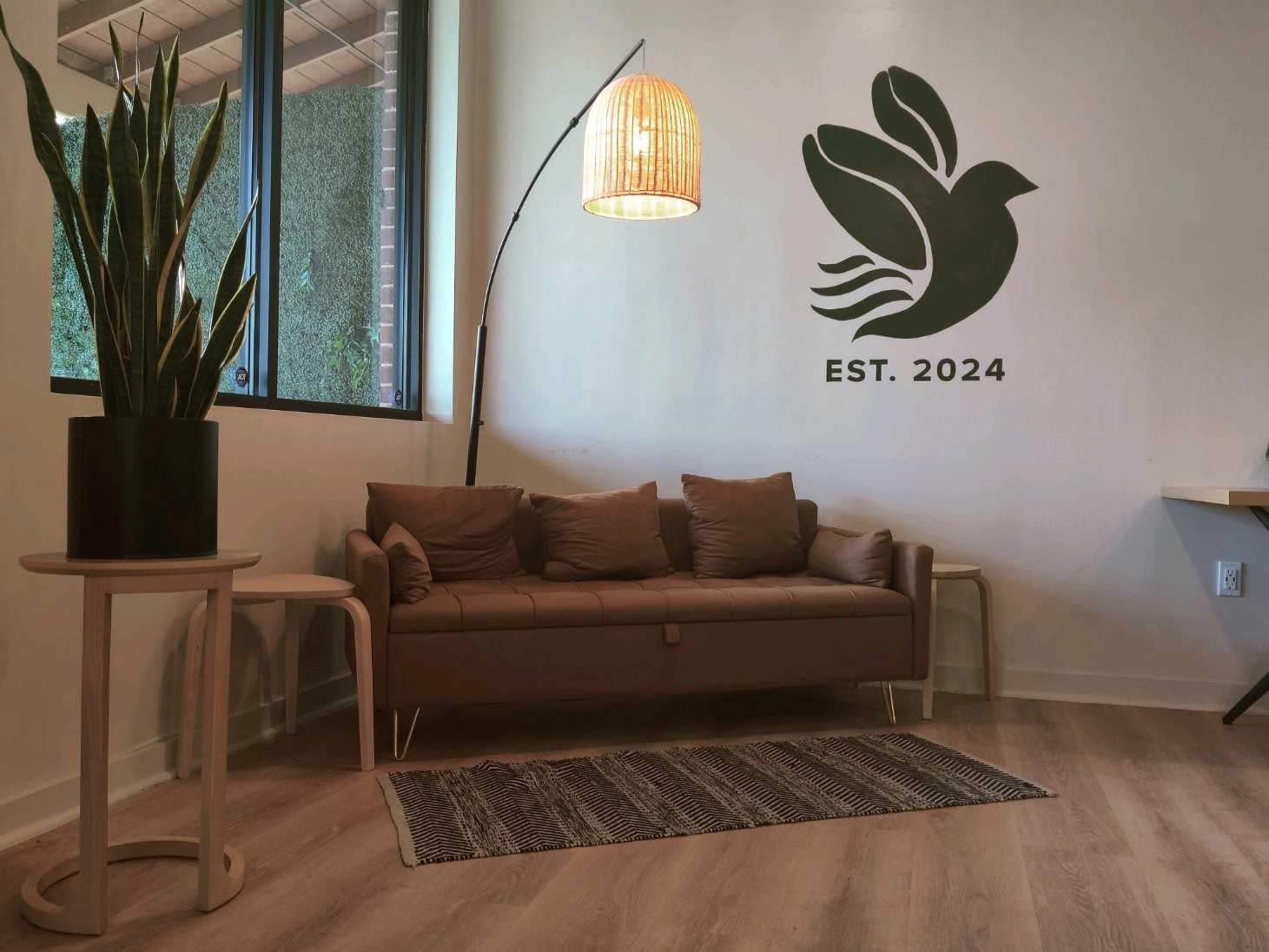 The image shows a modern indoor space featuring a brown sofa with cushions, a round side table, a tall plant in a black pot, and a decorative wall with a dove design and the text "EST. 2024."