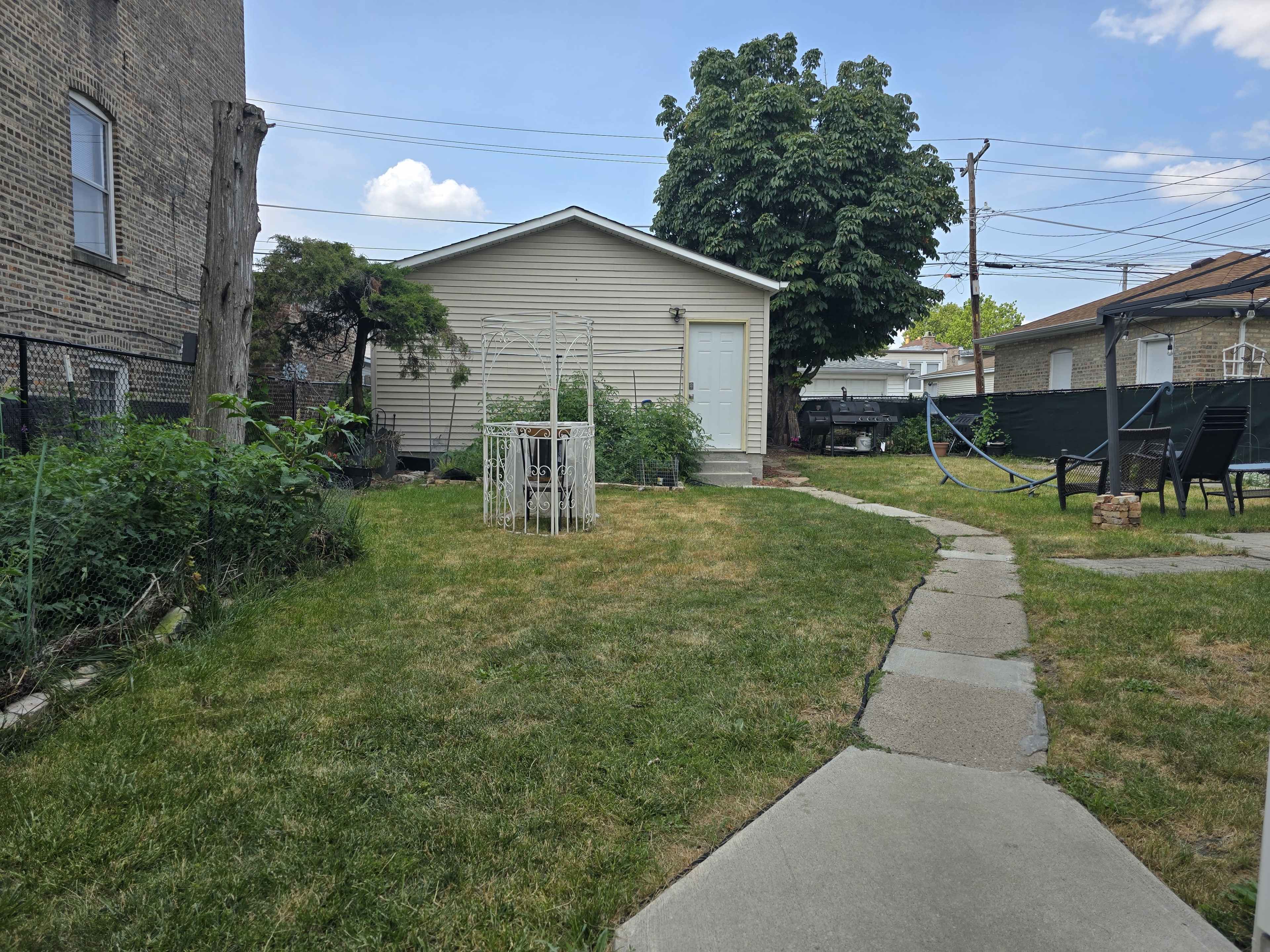 The image shows a backyard with a garden area, a small shed or house, and a stone pathway leading through the grass.
