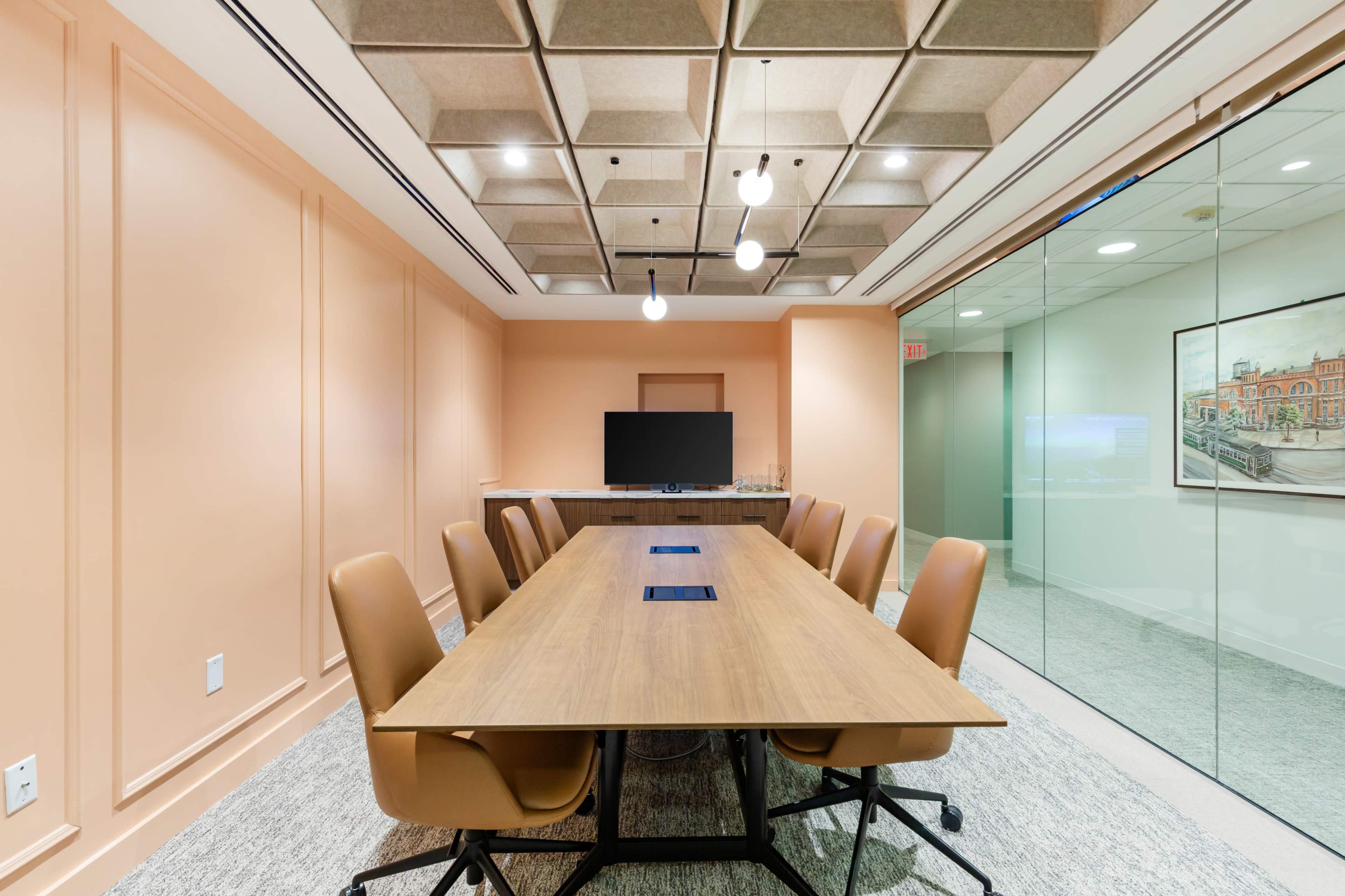 A modern conference room features a long wooden table surrounded by ergonomic chairs, with a television mounted on the wall and large glass panels allowing natural light.
