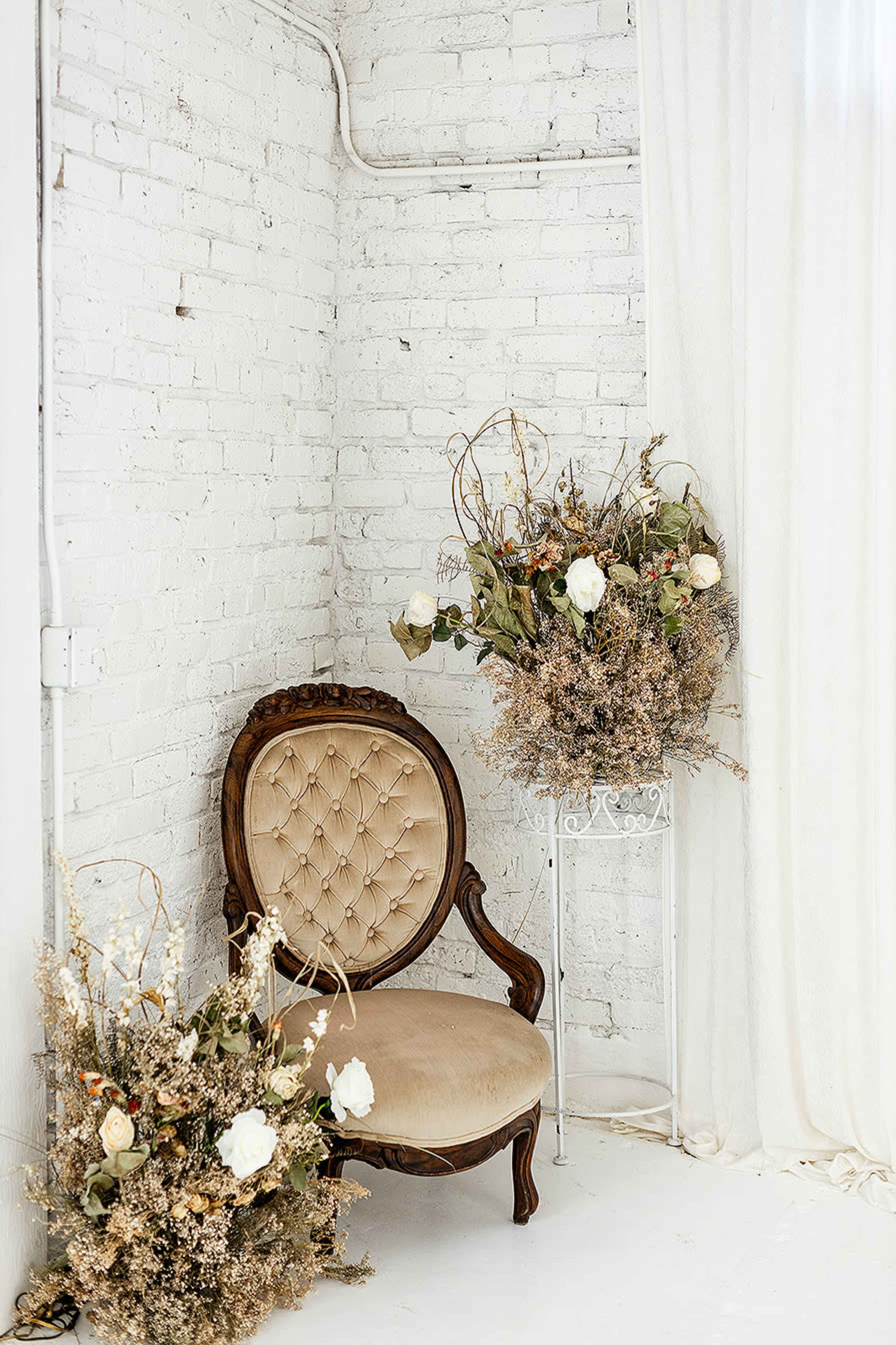 A vintage velvet chair is positioned against a white brick wall, accompanied by two large floral arrangements in neutral tones.