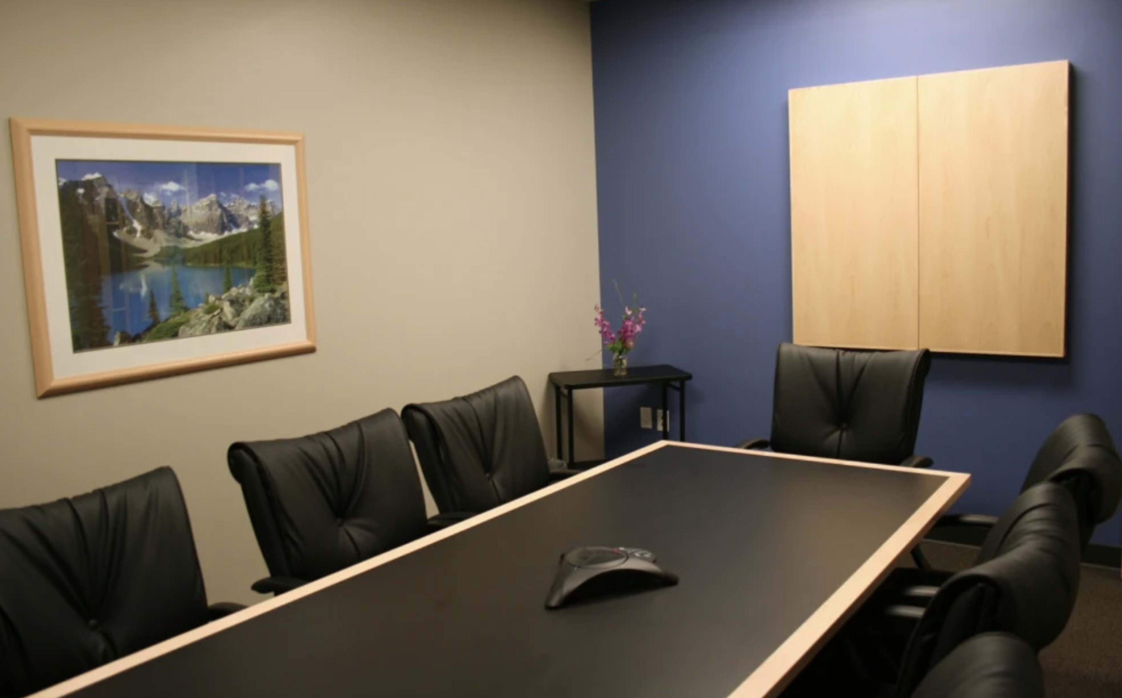 A conference room with a black table surrounded by black chairs, a framed landscape picture on one wall, and a wooden panel on another.