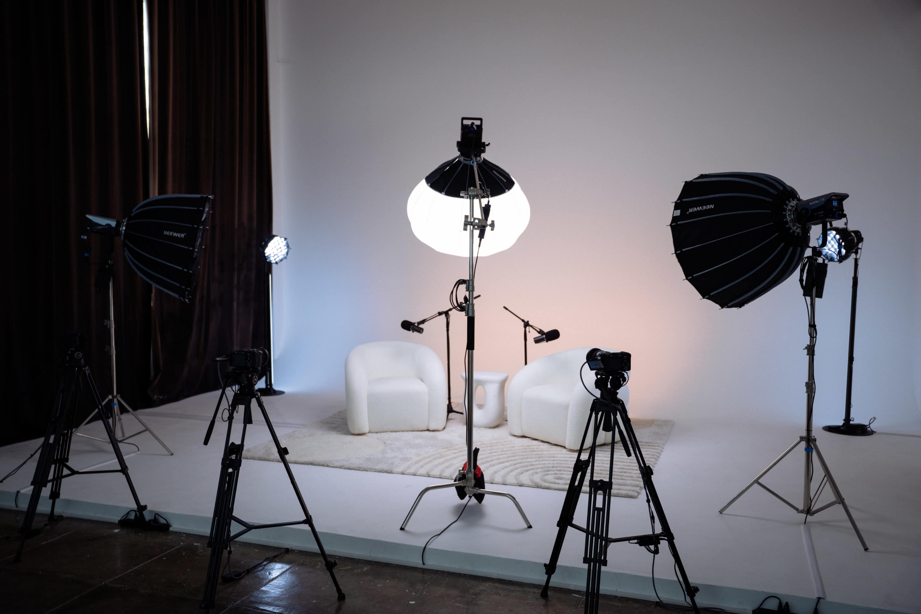 The image shows a photography studio setup with white chairs, lighting equipment, and cameras arranged around a neutral backdrop.