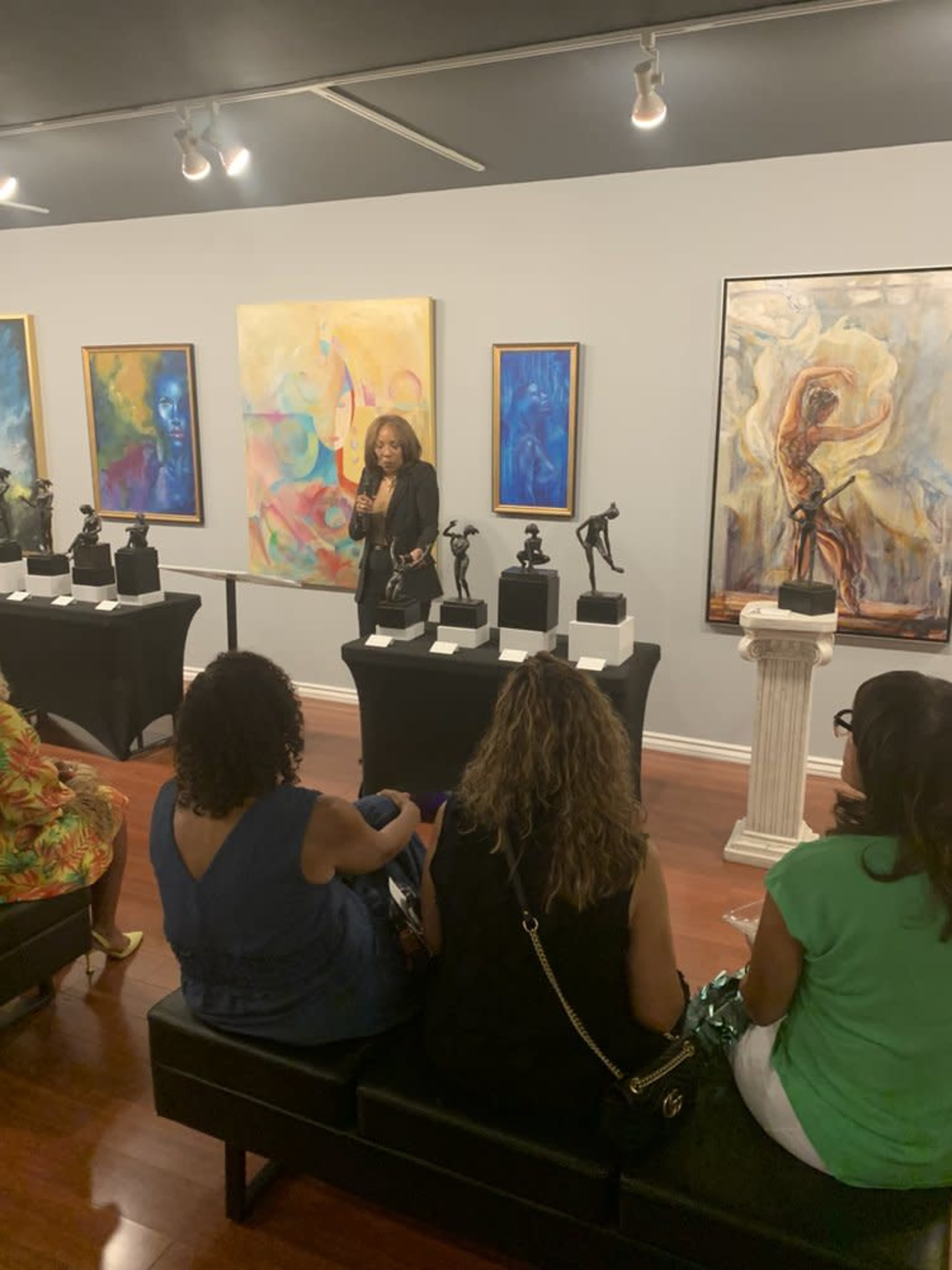 A speaker addresses an audience in an art gallery, surrounded by sculptures and abstract paintings on the walls.