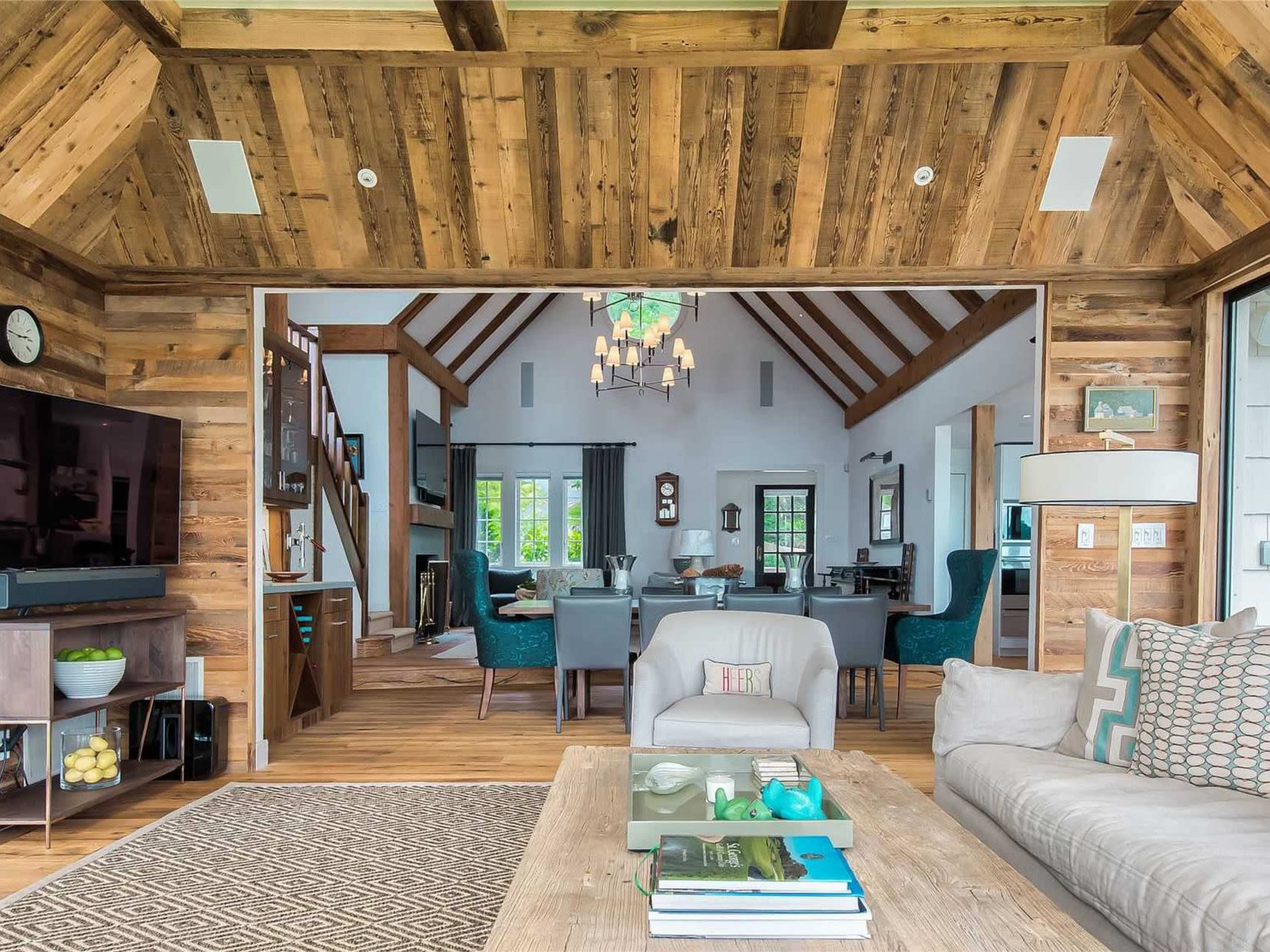 The image shows a rustic interior of a home featuring wooden beams, a living area with a sofa and coffee table, and an open space leading to a dining area.