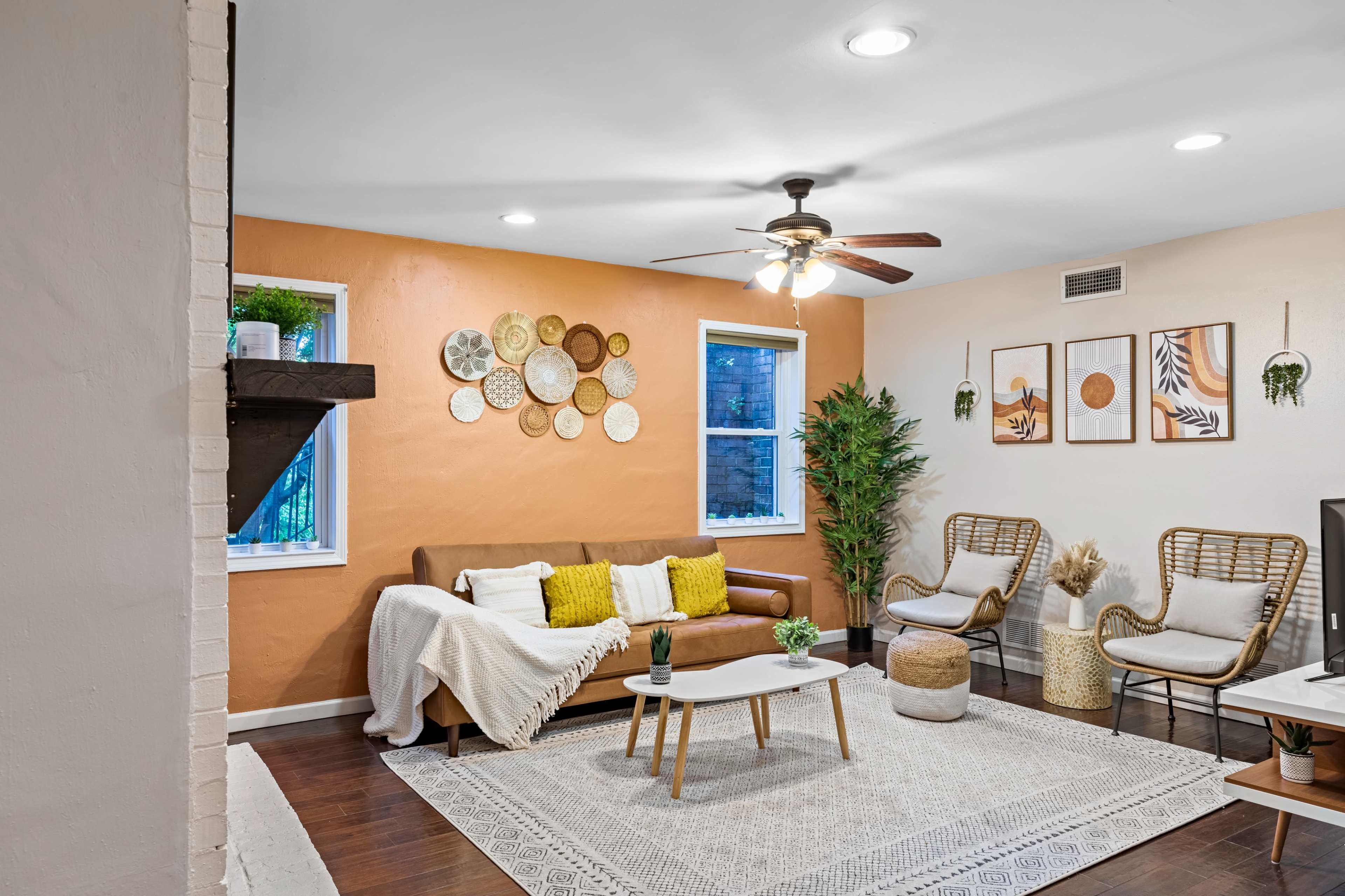The image shows a living room with a brown sofa, a coffee table, two wicker chairs, and decorative wall art against an orange accent wall.