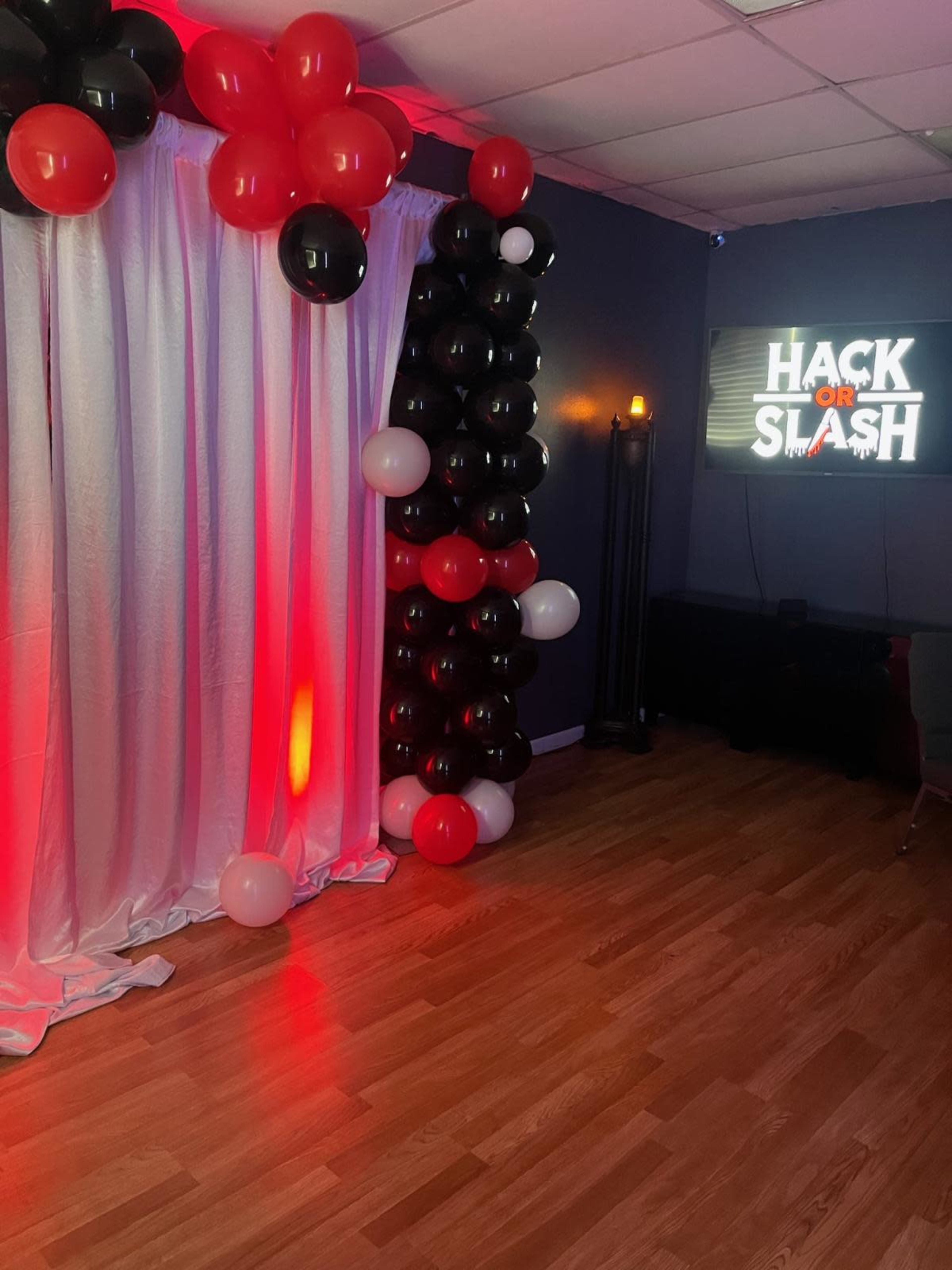 The image shows a corner of a room decorated with a white curtain, a balloon arch in black, white, and red colors, and a sign that reads "HACK OR SLASH."