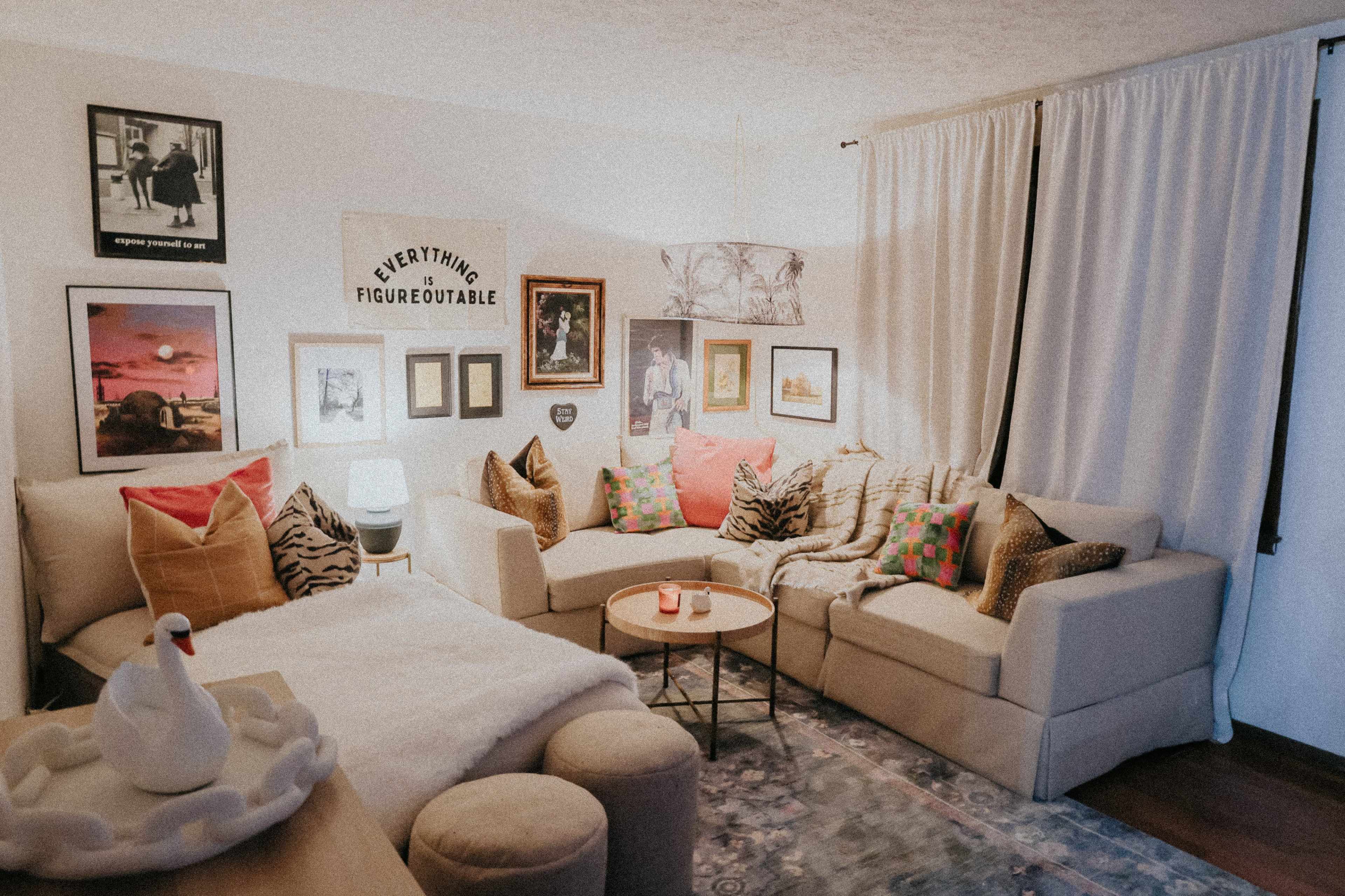 The image shows a cozy living room featuring a light-colored sectional sofa adorned with various decorative pillows, framed artwork on the walls, and a coffee table with a small decorative item.