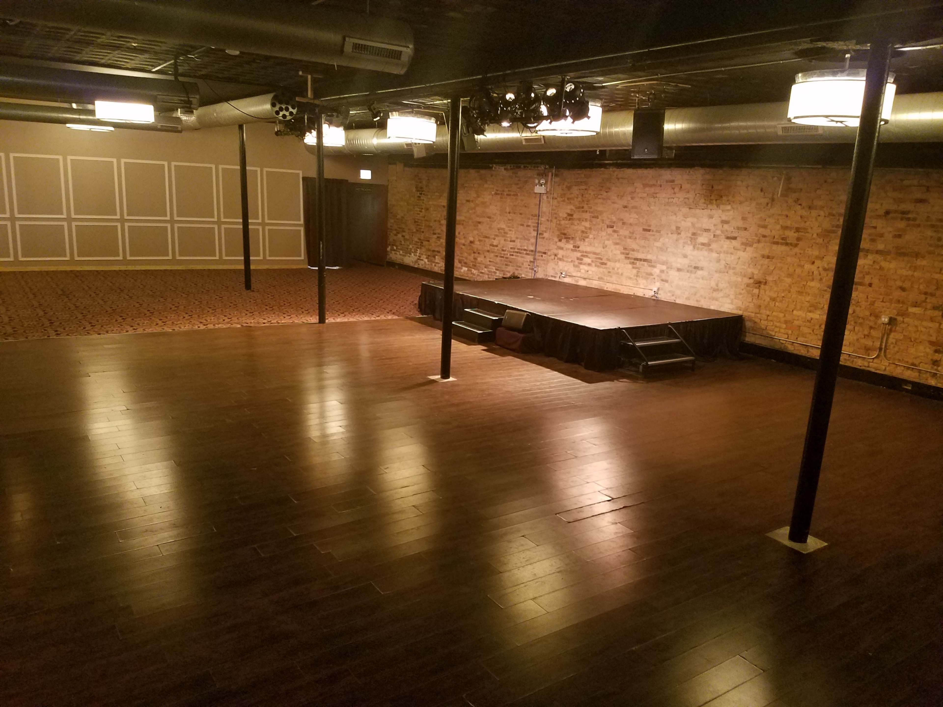 An empty event space featuring a wooden floor, exposed brick walls, a small stage, and overhead lighting fixtures.