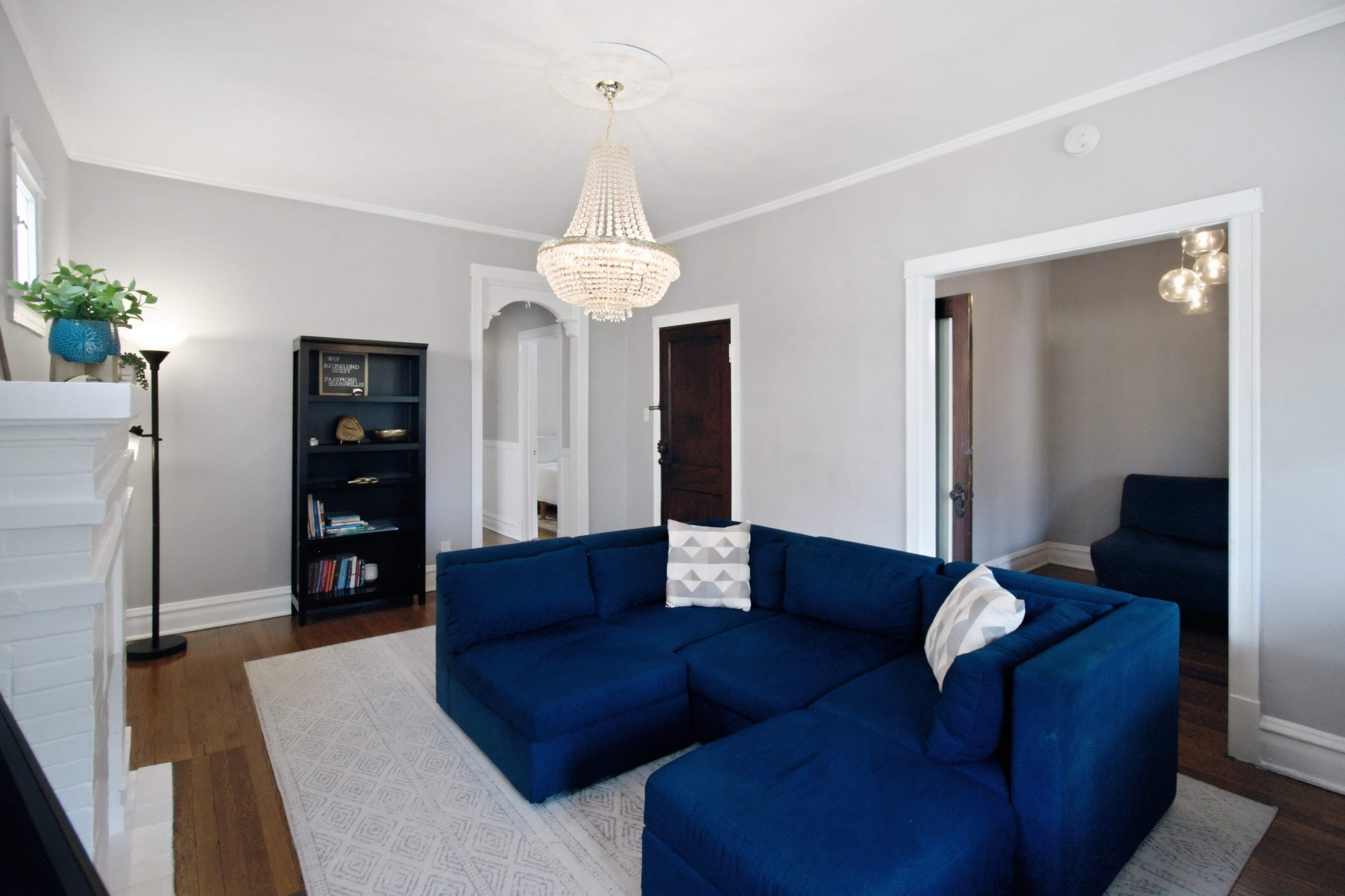 The image shows a modern living room featuring a blue sectional sofa, a chandelier, and a bookshelf against a gray wall.