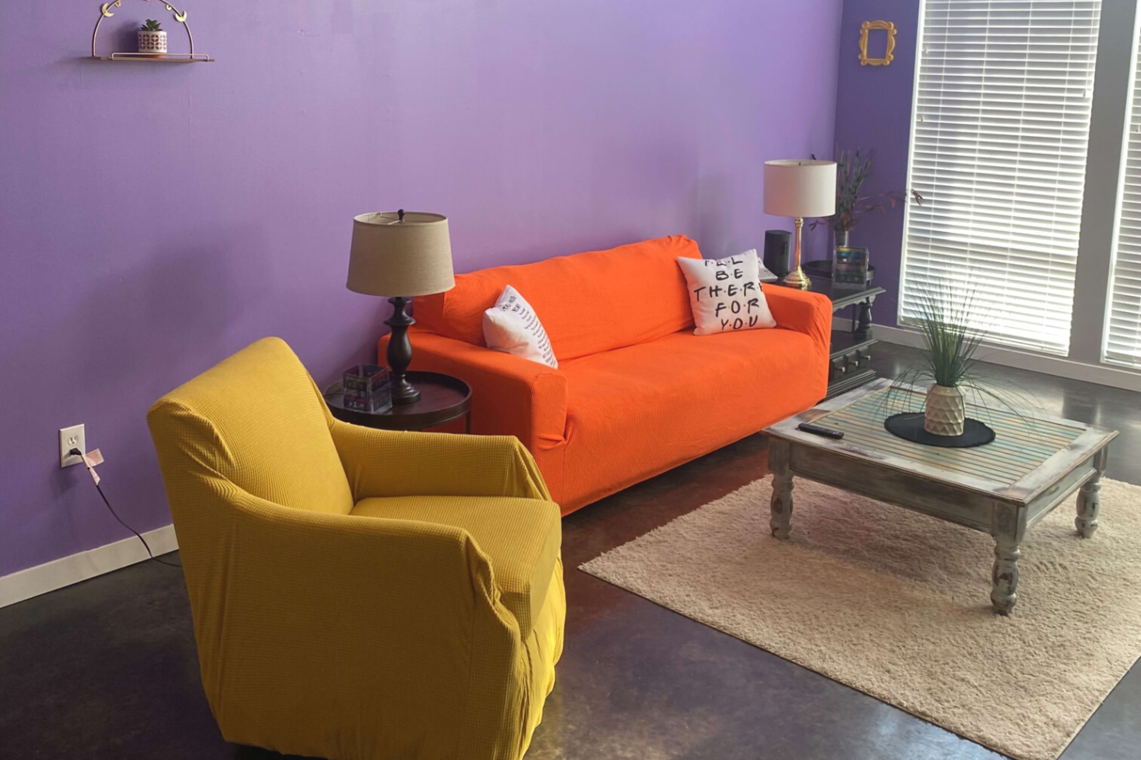 A cozy living room with a purple wall, an orange sofa, a yellow chair, a wooden coffee table, and two lamps beside the windows.