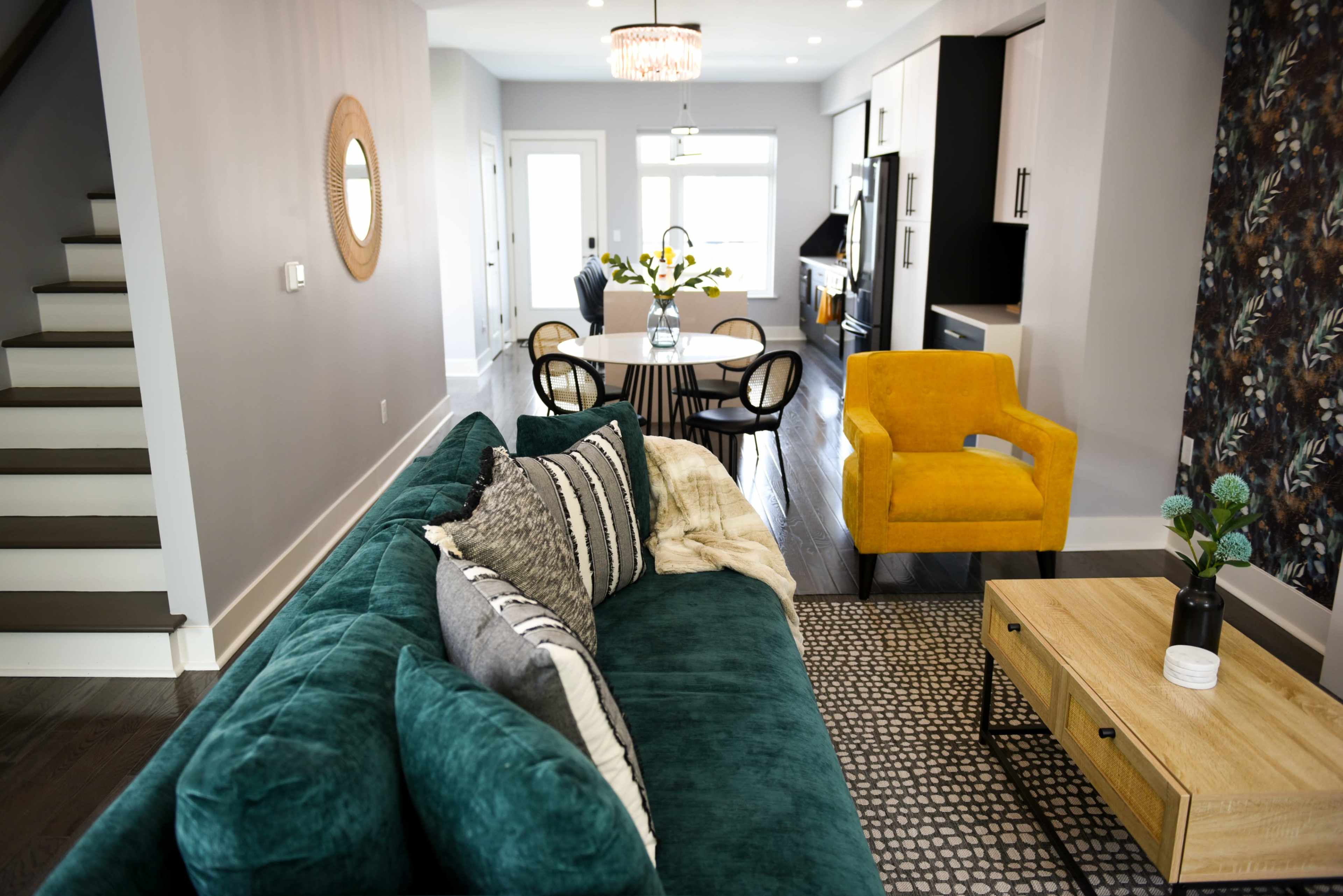A modern living space featuring a teal sofa, a yellow armchair, a dining table with chairs, and a kitchen area in the background.