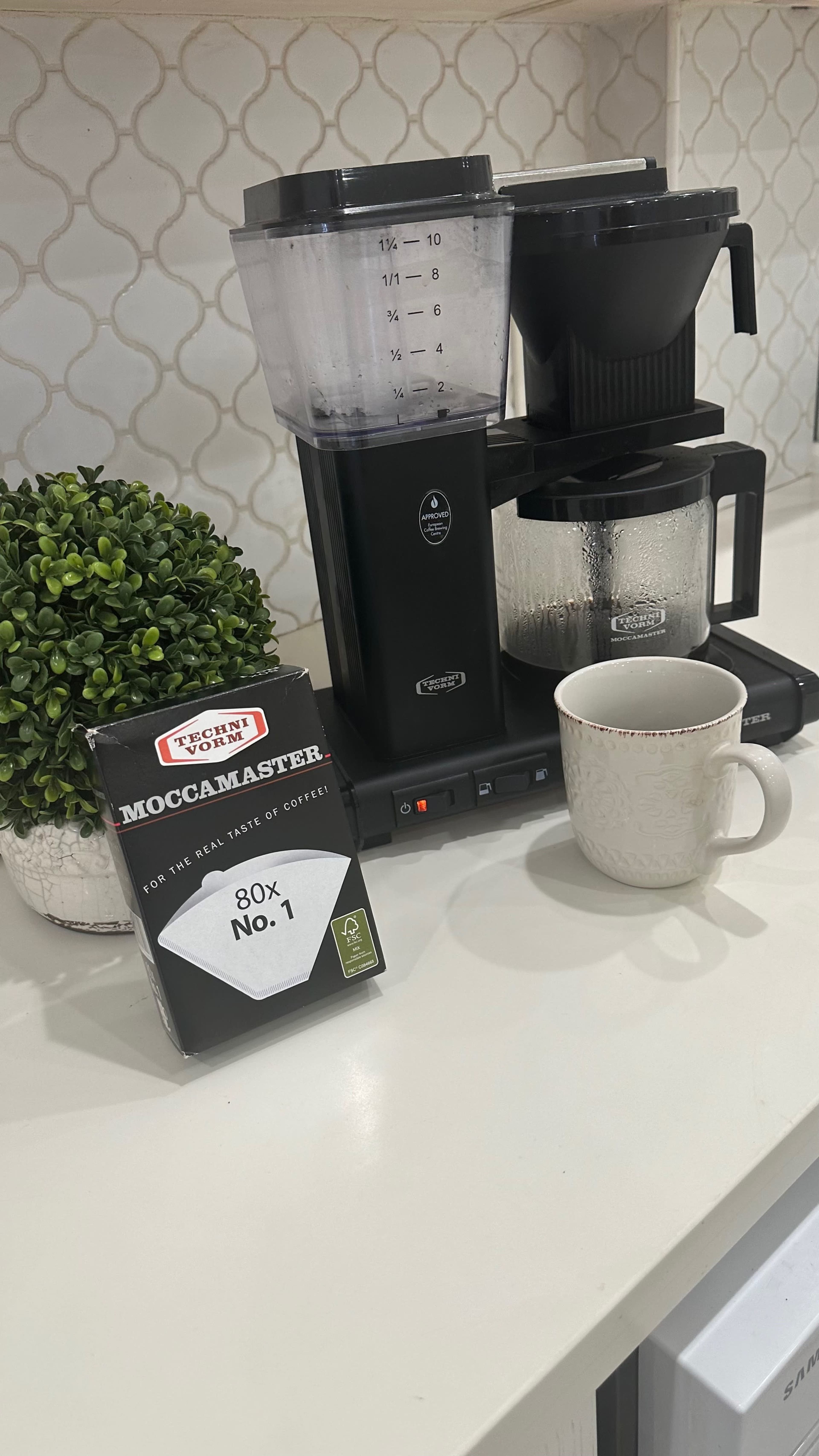 A Moccamaster coffee maker sits on a countertop next to a box of coffee filters, a mug, and a decorative potted plant.