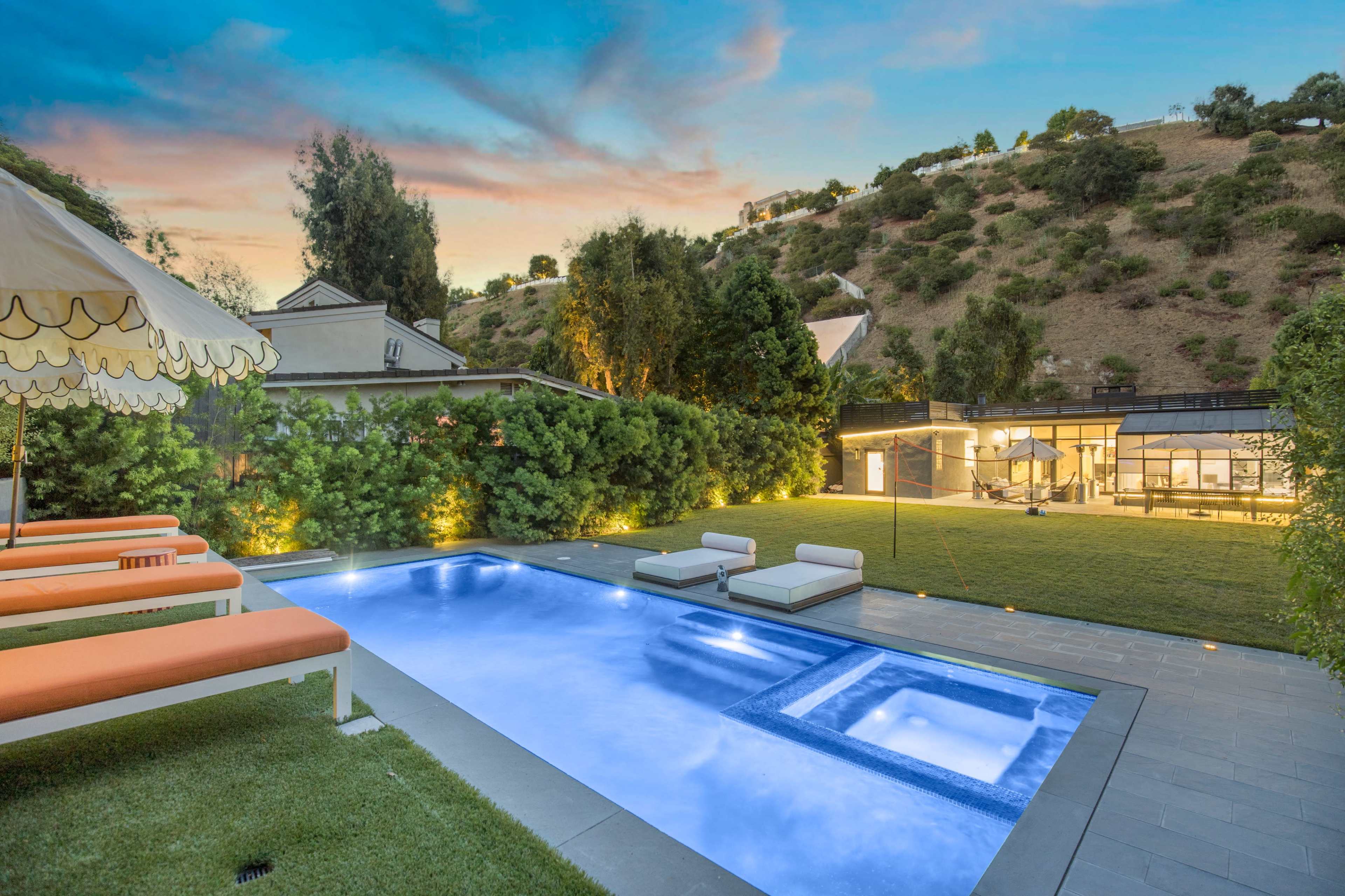 The image shows a modern backyard featuring a swimming pool, lounge chairs, and a neatly landscaped lawn, with a hillside in the background.