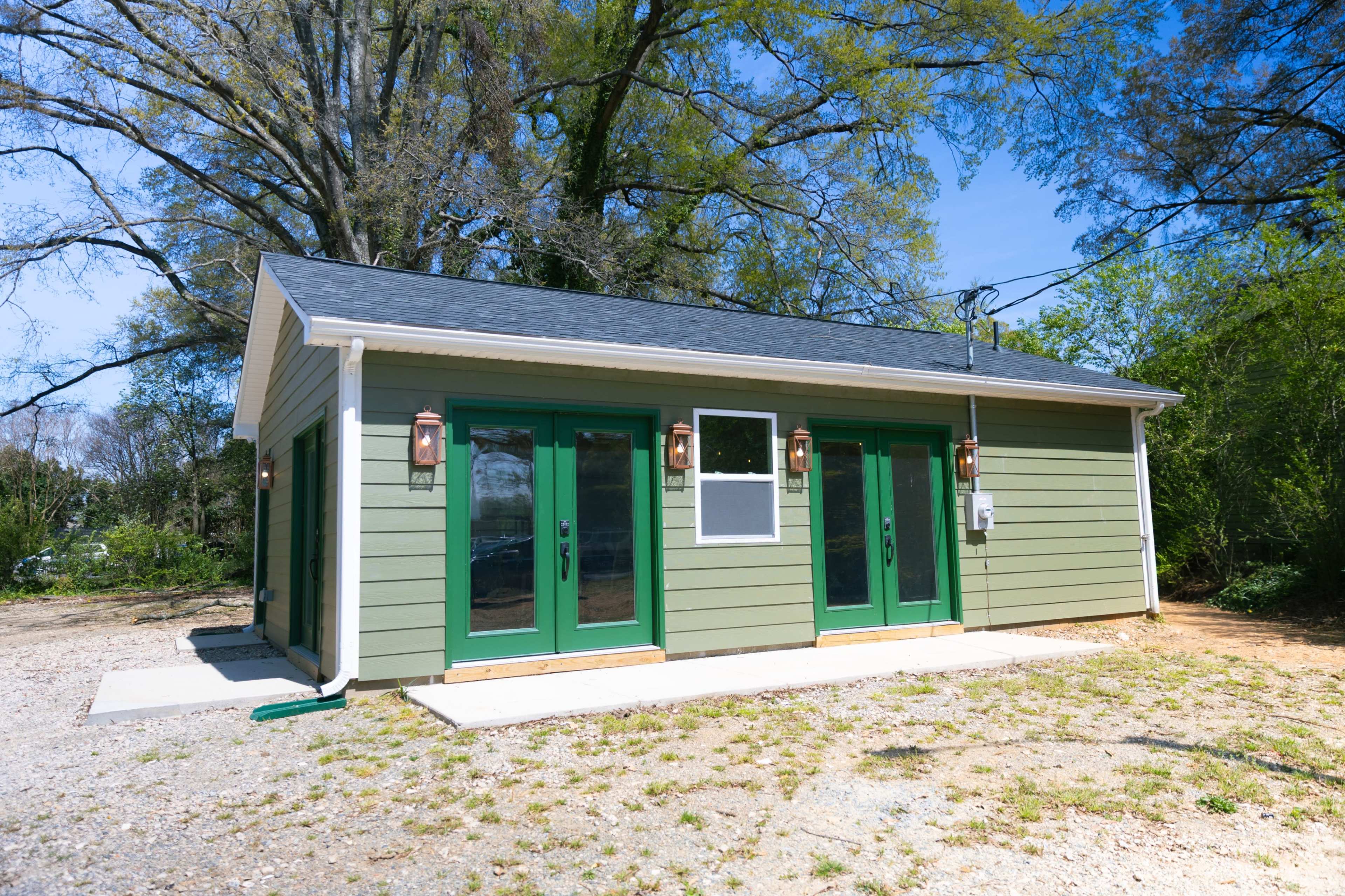 The image shows a small green building with multiple green doors and windows, situated in a gravel area with trees in the background.