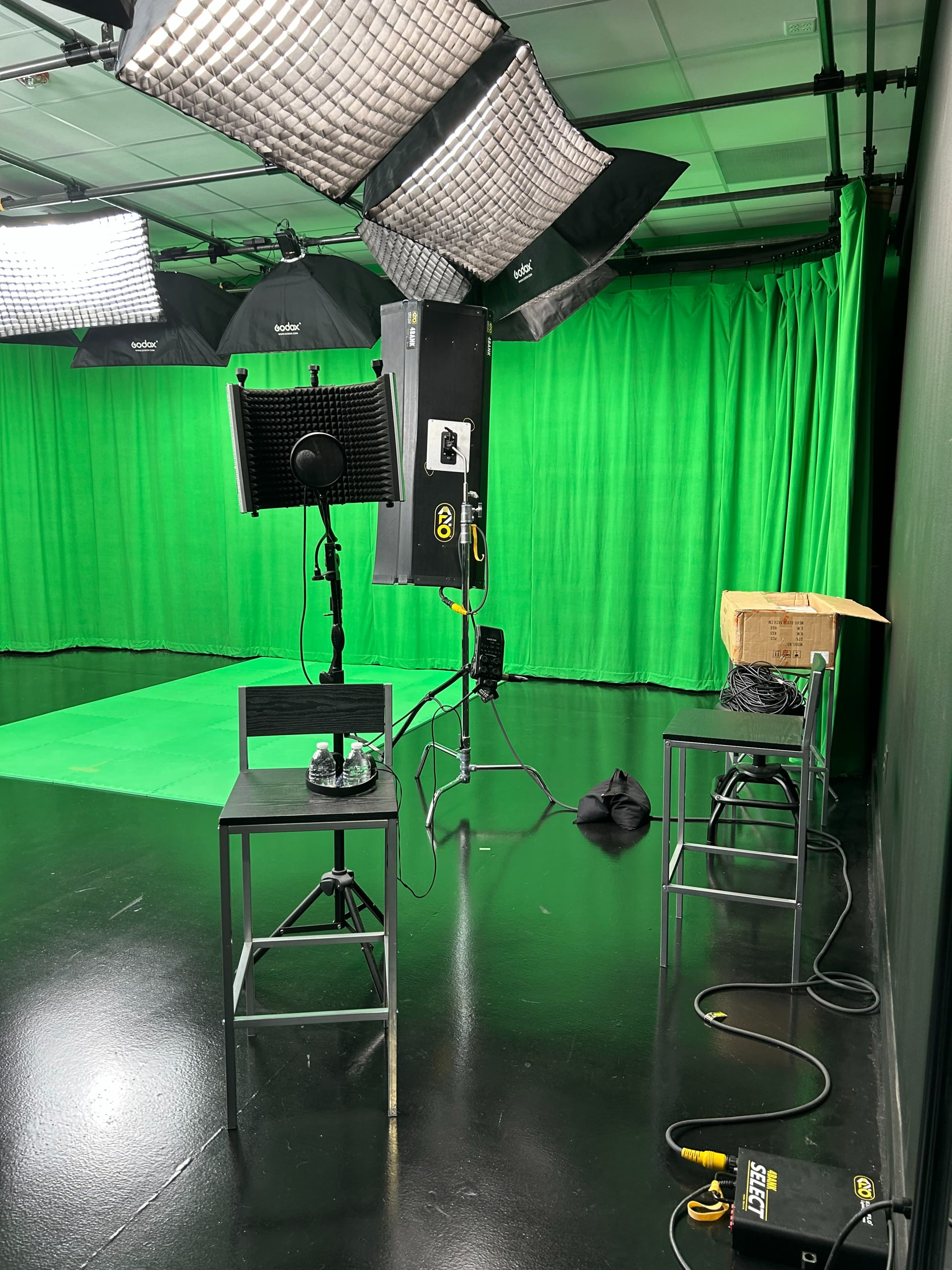 The image shows a green screen studio setup with lighting equipment, two empty chairs, and cables on a glossy black floor.