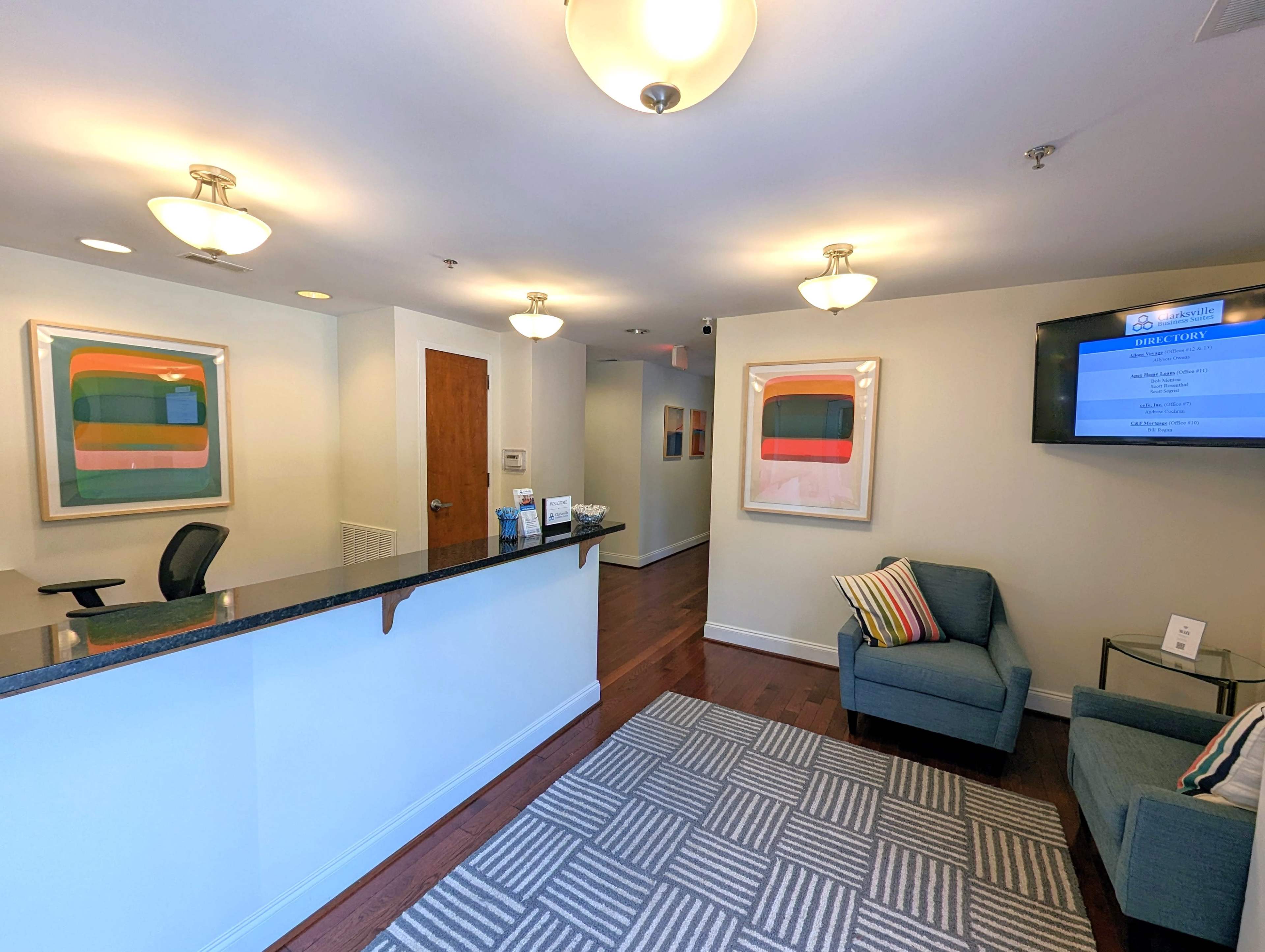 The image shows a reception area with a desk, two chairs, framed artwork on the walls, and a television displaying information.