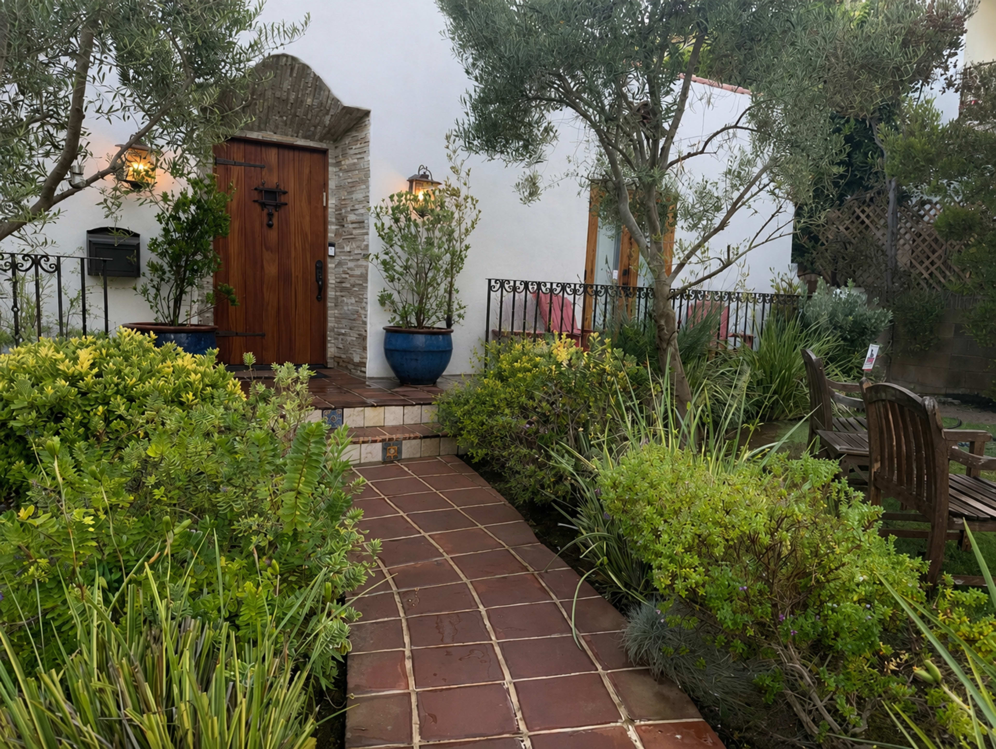 A brick pathway leads to a wooden door framed by potted plants and a small seating area in a lush garden.