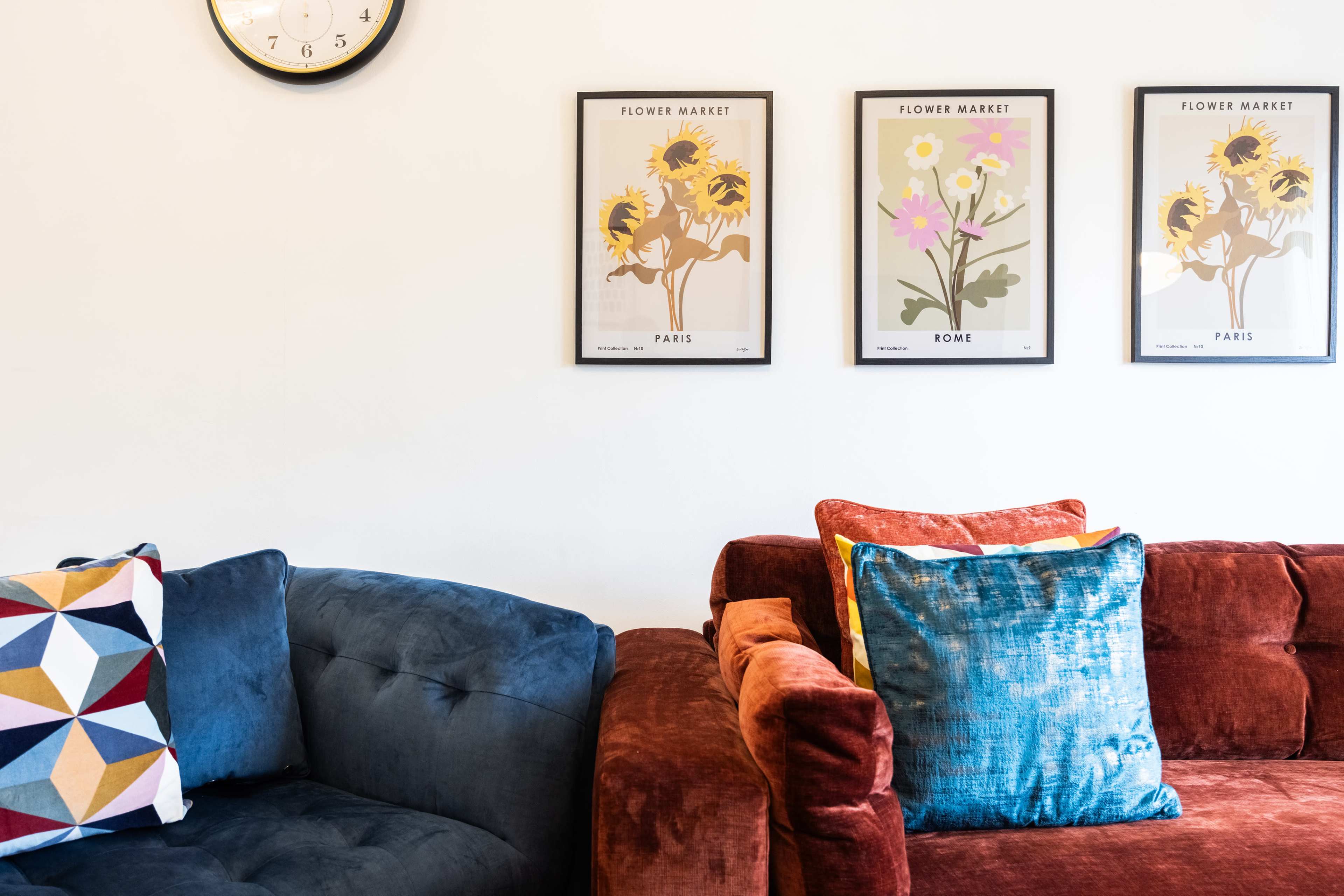 Two distinct sofas, one blue and one orange, situated below three flower market posters and accompanied by a wall clock.