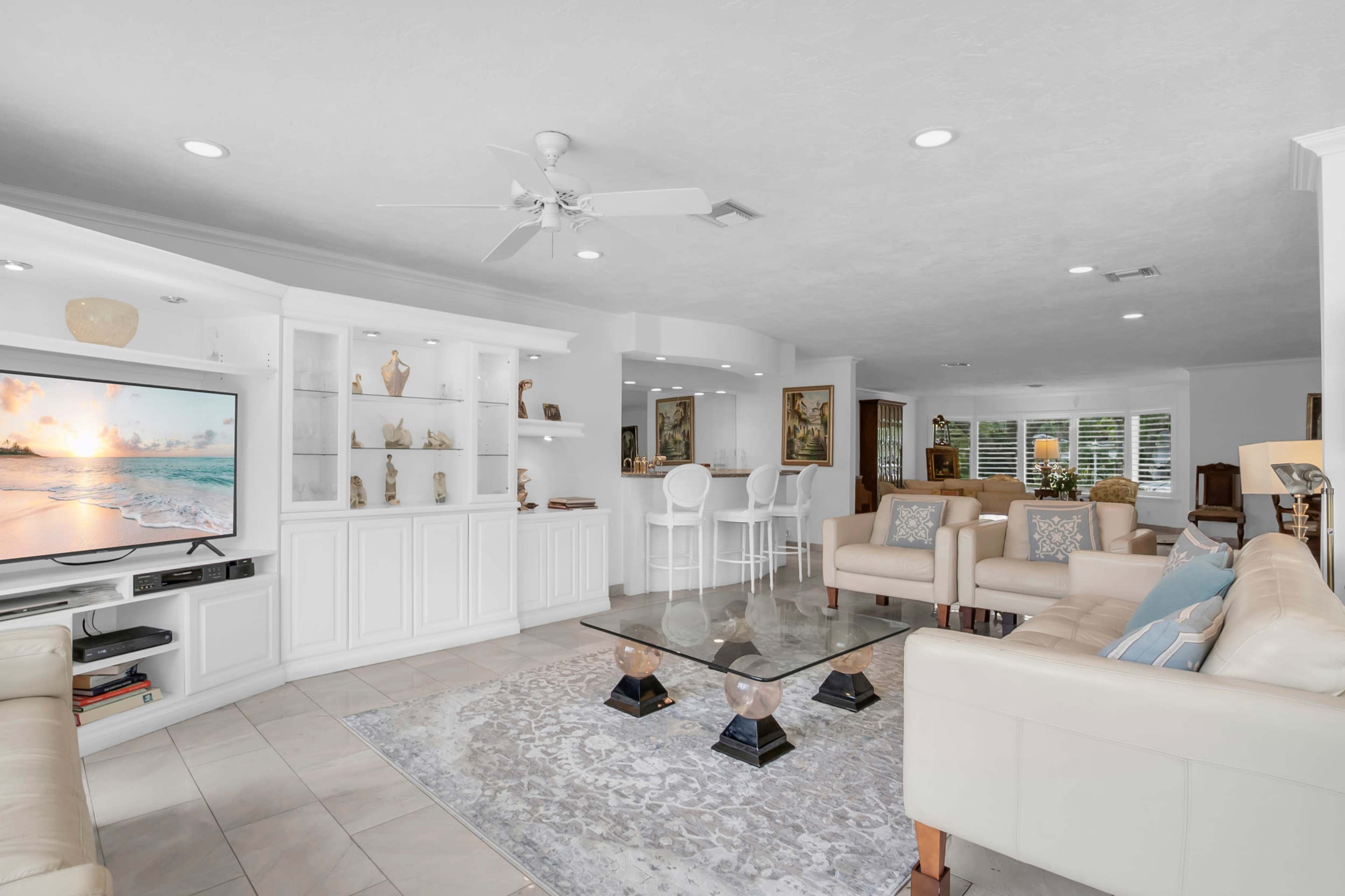 The image shows a modern living room with a white entertainment unit, beige sofas, a light rug, and a glass coffee table, accompanied by a dining area in the background.