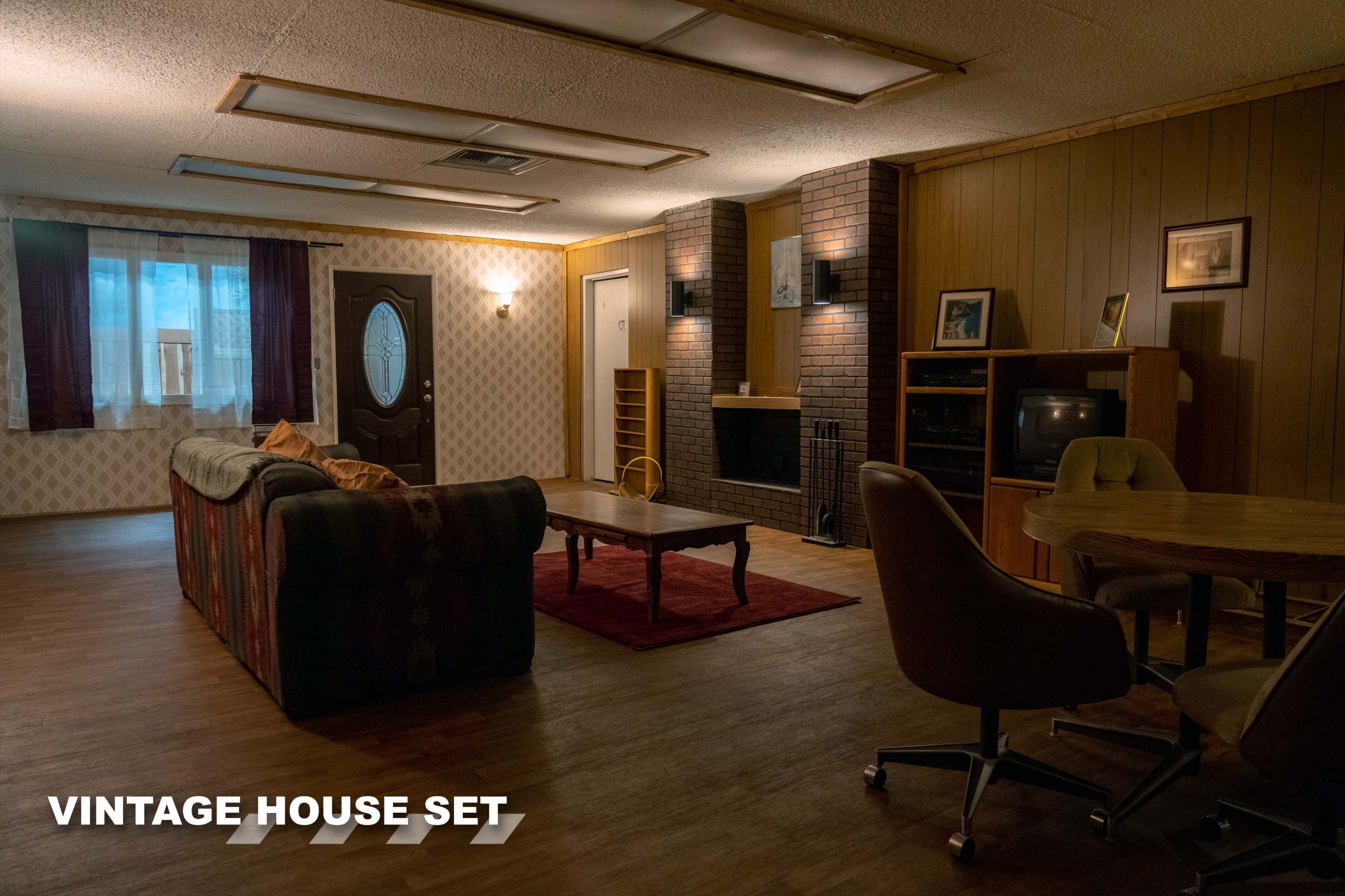 The image depicts a vintage house set featuring a living area with a sofa, coffee table, dining table, and a brick fireplace against a textured wall.