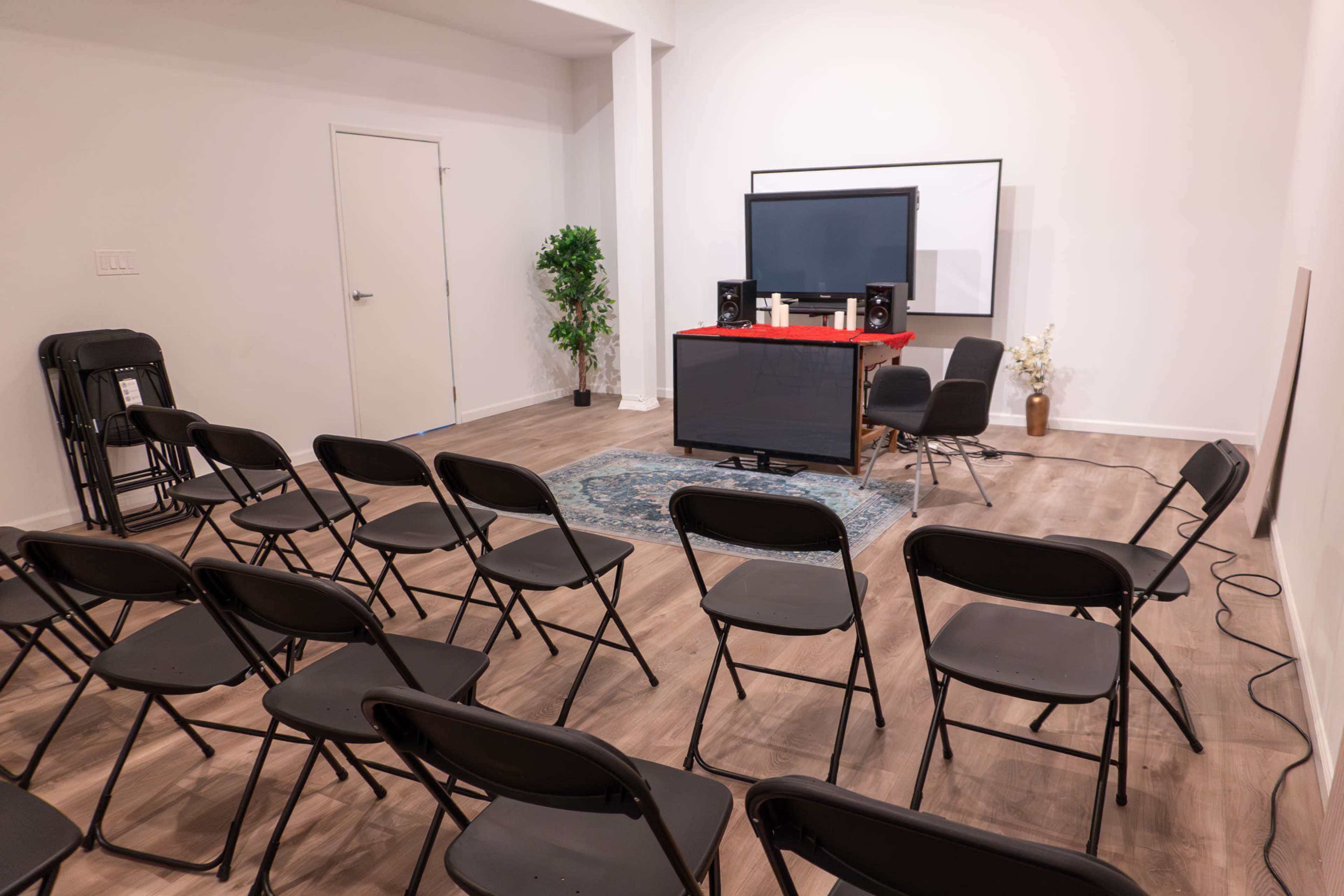 The image shows a room arranged with black folding chairs facing a table with speakers and a large screen at the front, complemented by a plant in the corner.