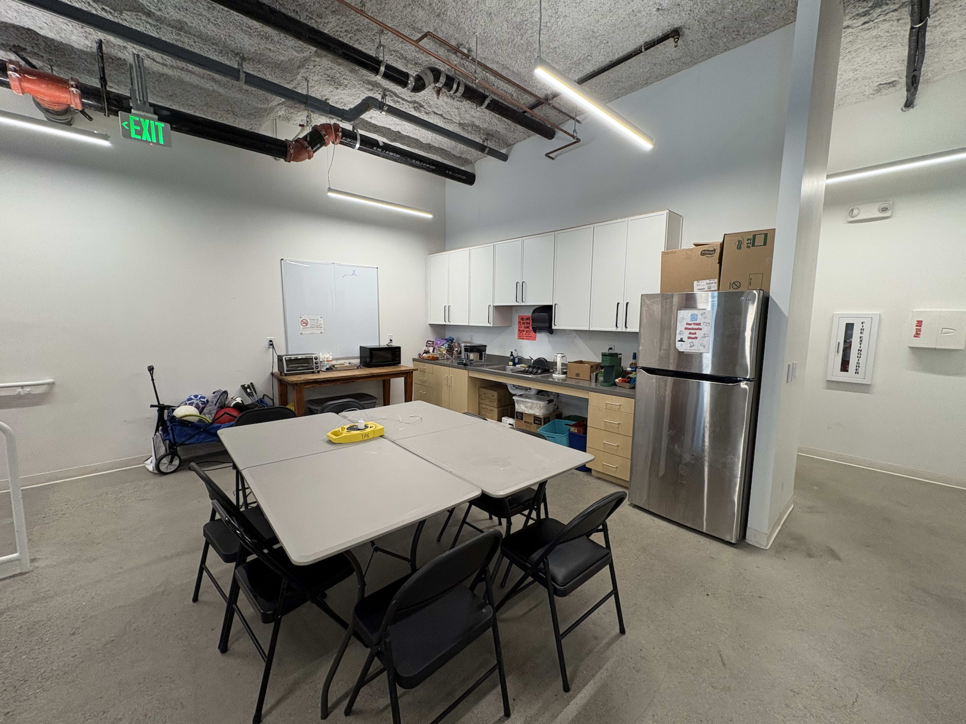 The image shows a clean kitchen area with a table and chairs, a refrigerator, and various kitchen appliances and supplies on shelves and counters.