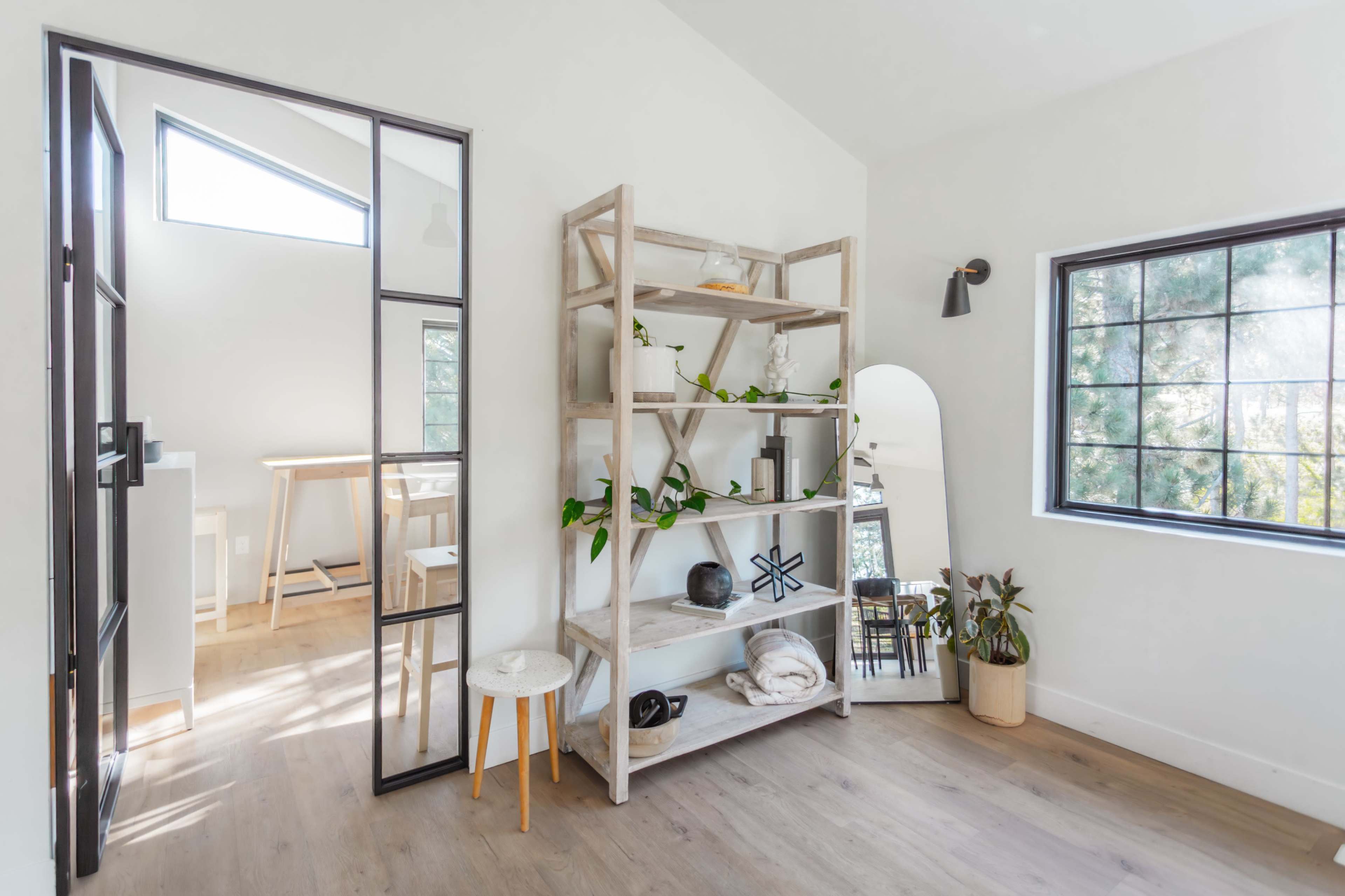 A bright room featuring a tall shelving unit, a round stool, a large mirror, and a partially open door leading to another space.
