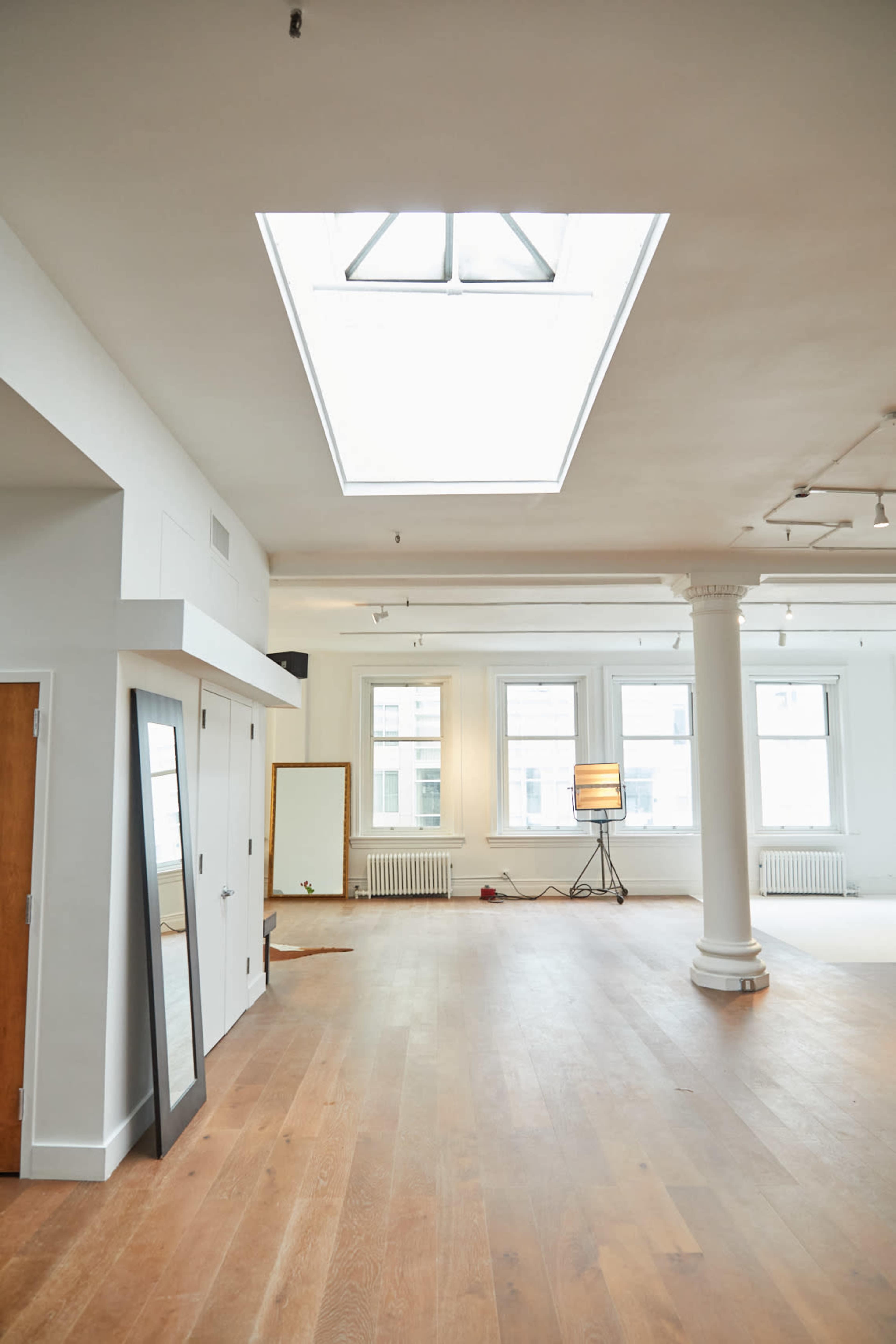 The image shows a spacious, well-lit room with wooden floors, featuring large windows, a skylight, and a floor lamp, along with a full-length mirror against one wall.