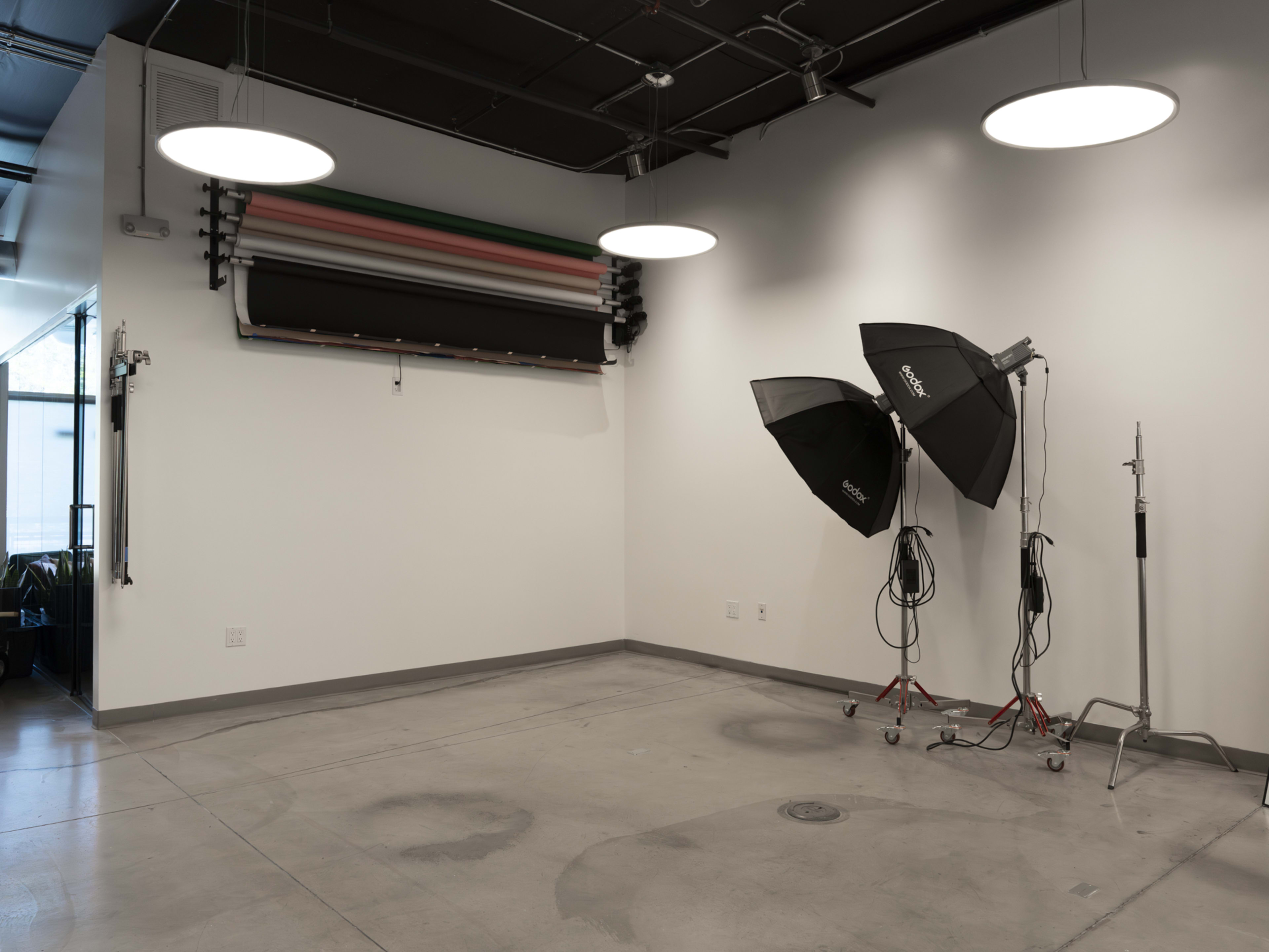 The image shows a studio space with a plain wall, backdrop rolls, and two softbox lighting setups on stands.